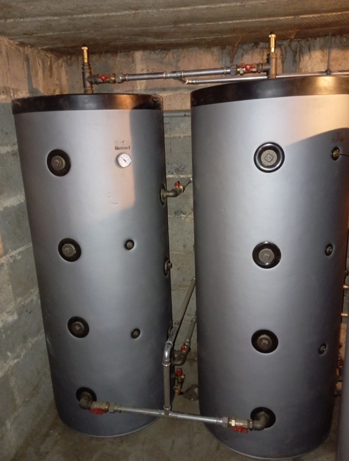 Dwa szare, cylindryczne zbiorniki z widocznymi przyłączami rur i zaworami, umieszczone w piwnicznym pomieszczeniu z betonową ścianą i podłogą.