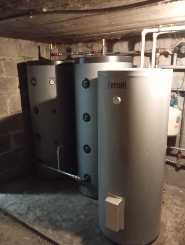 Trzy szare zasobniki ciepłej wody użytkowej marki Ferroli w piwnicy, widoczne rury i armatura, surowy betonowy strop.