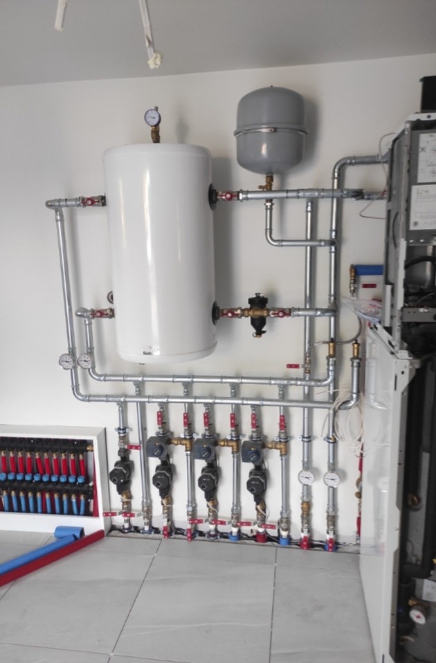 Kompleksowa instalacja grzewcza z białym zasobnikiem ciepłej wody, szarym naczyniem wzbiorczym i rozdzielaczem ogrzewania podłogowego z czerwono-niebieskimi pętlami, widoczna na tle białej ściany...