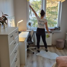 AGNIESZKA FIEDOSIUK CLEARITY - Kobieta w koszulce z napisem 'Wszystko w porządku' czyści pajęczynę pod sufitem w pokoju za pomocą żółtej zmiotki, widok z tyłu na tle okna, biurka i łóżka.