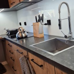 AGNIESZKA FIEDOSIUK CLEARITY - Nowoczesna kuchnia z drewnianymi szafkami, stalowym zlewem, czajnikiem i listwą nożową na blacie, z oświetleniem LED pod szafkami górnymi.