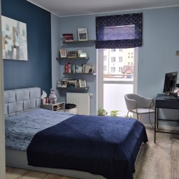 AGNIESZKA FIEDOSIUK CLEARITY - Uporządkowana sypialnia w odcieniach szarości i granatu, z łóżkiem z szarym zagłówkiem i granatową narzutą, drewnianą podłogą, biurkiem z komputerem i półkami z książkami.