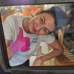 AGNIESZKA FIEDOSIUK CLEARITY - Uśmiechnięta kobieta w różowych rękawicach, ubrana w białą koszulkę z logo firmy sprzątającej, daje kciuk w górę, patrząc przez otwarte drzwiczki pieca kominkowego, widoczne wnętrze kuchni...