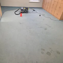 Szaro-niebieski dywan biurowy z widocznymi plamami, czyszczony za pomocą odkurzacza piorącego Karcher z czerwonym spryskiwaczem obok, w tle rząd szafek pracowniczych.