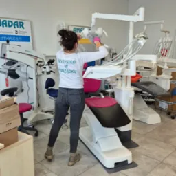 Kobieta w koszulce z napisem 'Wszystko w porządku' czyści za pomocą miotełki do kurzu biały unit stomatologiczny w gabinecie dentystycznym.