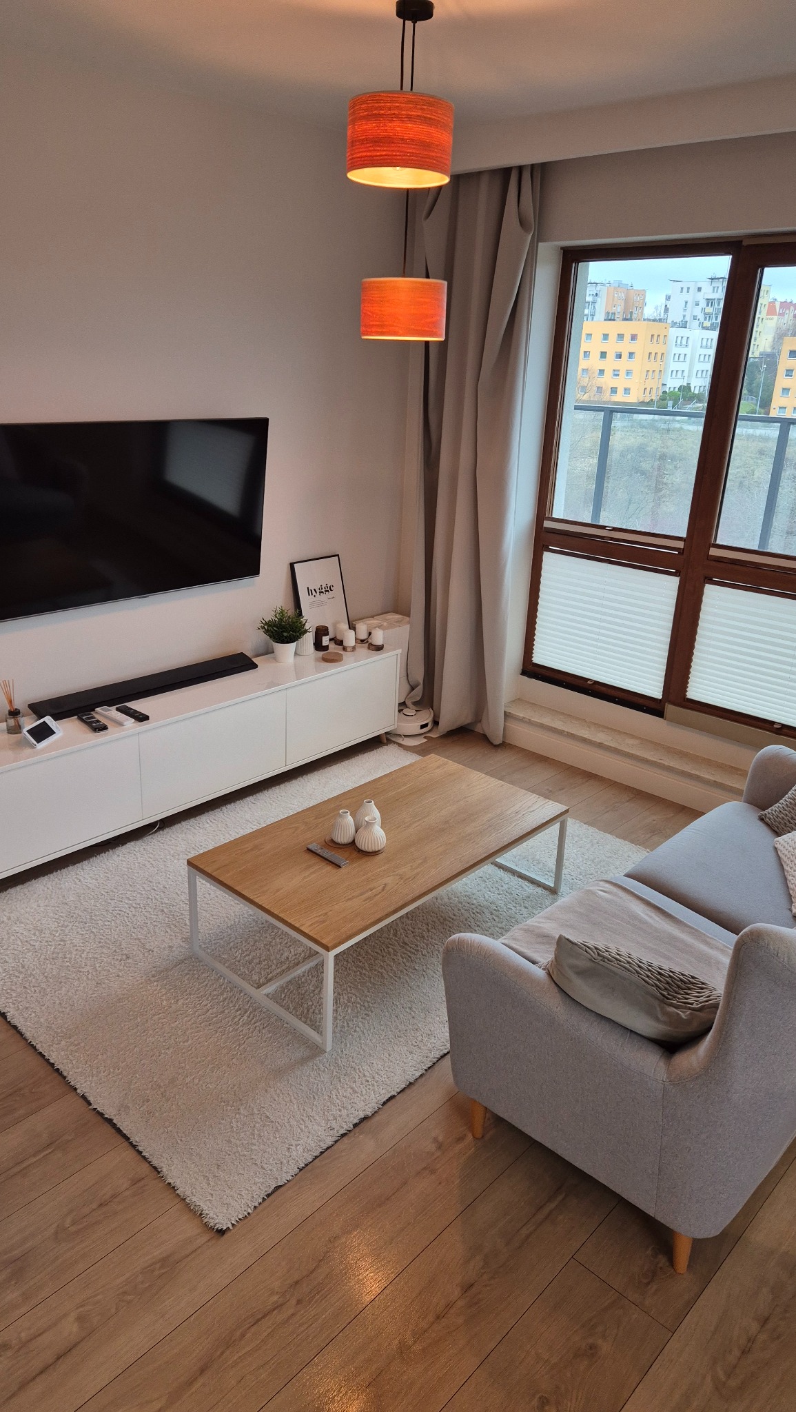 Jasne, nowocześnie urządzone wnętrze salonu z drewnianą podłogą, puszystym dywanem, szarą sofą i stolikiem kawowym; widok na osiedle zza okna z roletami plisowanymi.