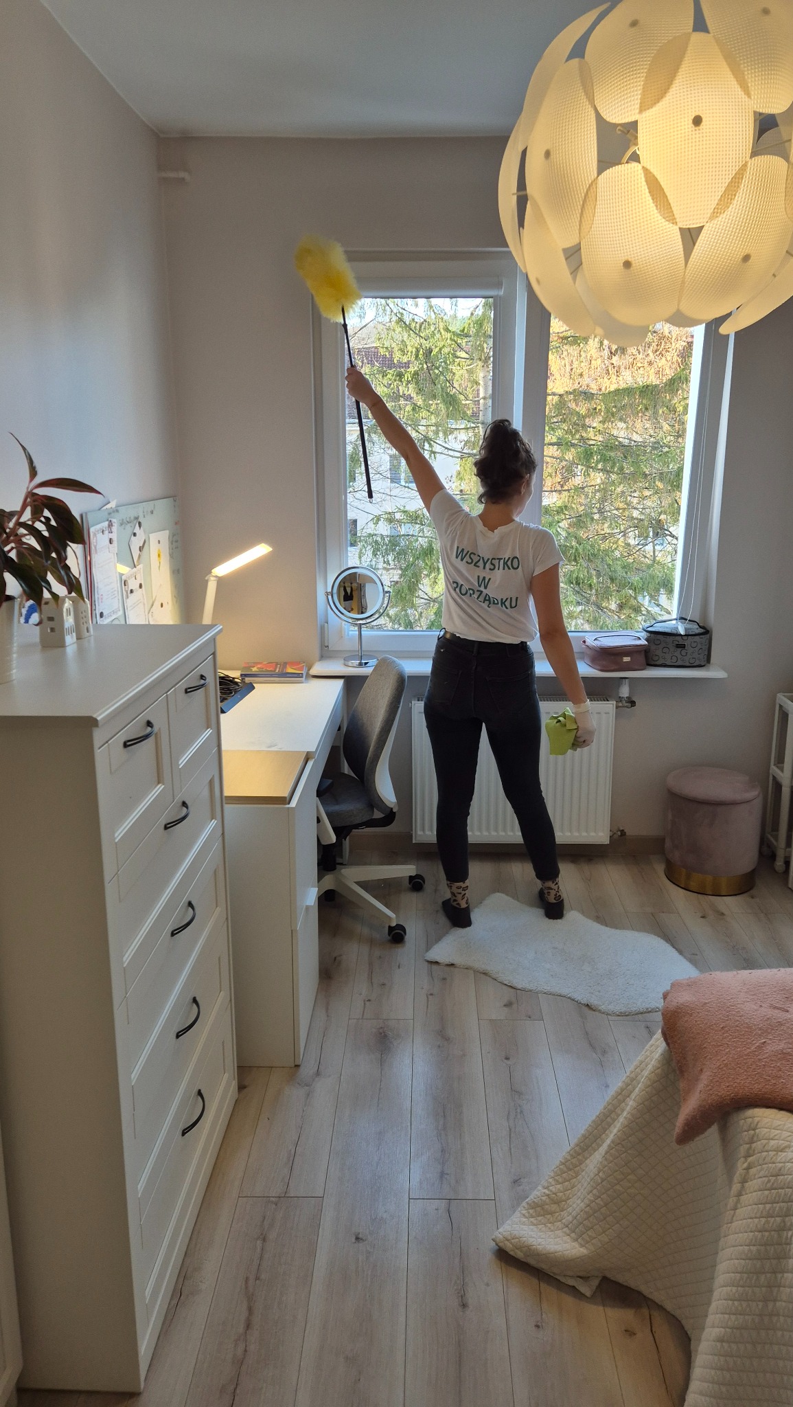 Kobieta w koszulce z napisem 'Wszystko w porządku' czyści pajęczynę pod sufitem w pokoju za pomocą żółtej zmiotki, widok z tyłu na tle okna, biurka i łóżka.