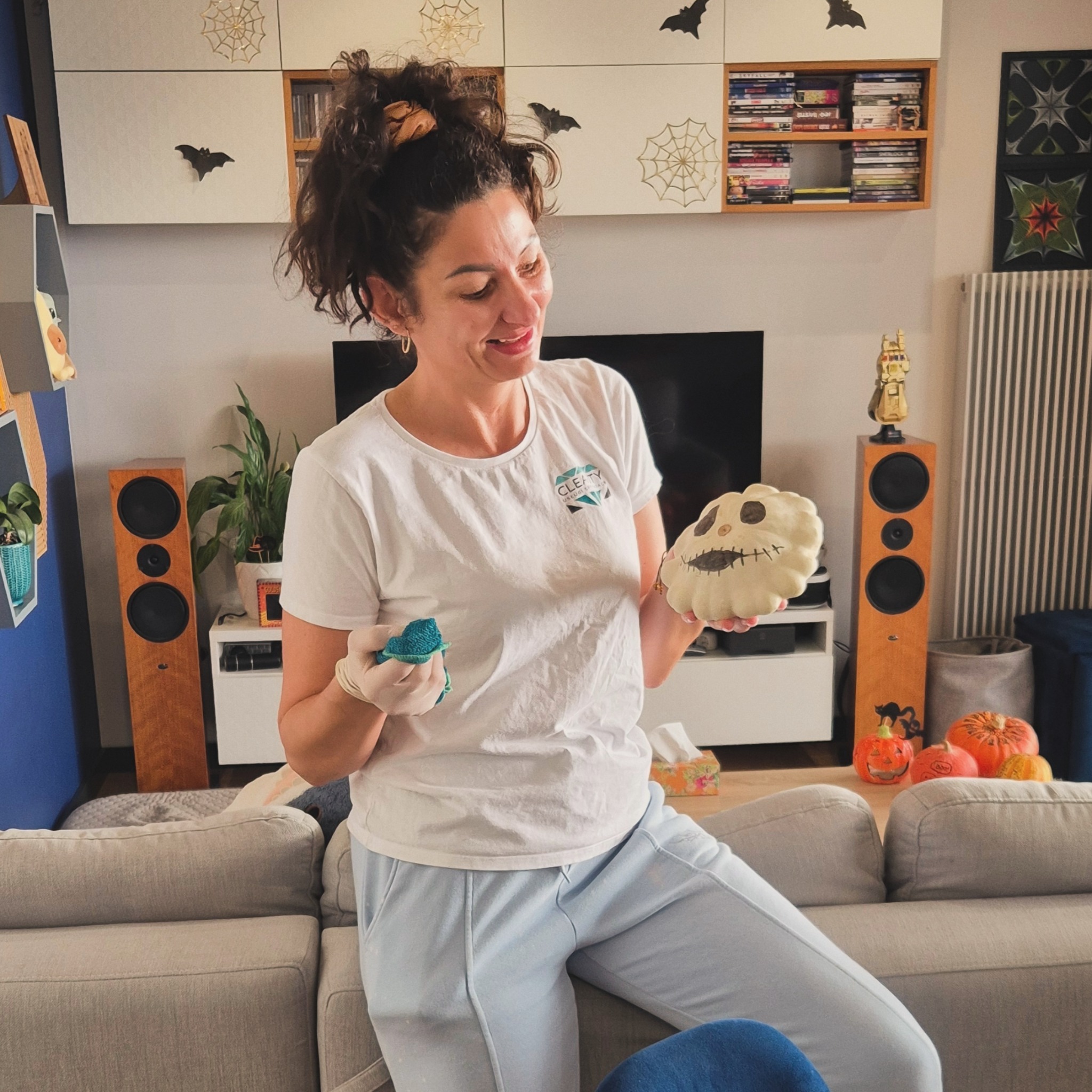 Uśmiechnięta kobieta w białej koszulce z logo firmy trzyma w dłoniach ścierkę i dekoracyjną dynię z wyciętą twarzą, stojąc w salonie udekorowanym na Halloween.