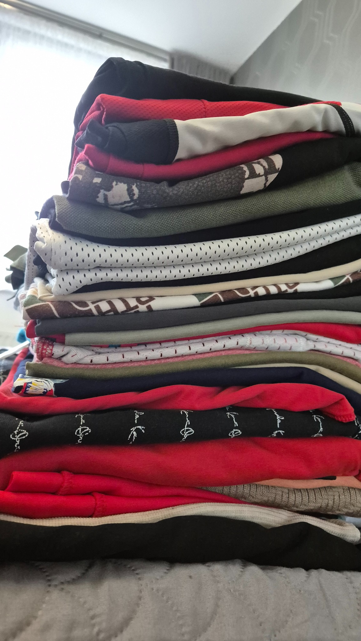 Stos świeżo złożonych ubrań, w tym koszulek i bluz, z różnymi wzorami i kolorami, leżący na szarej, pikowanej powierzchni.