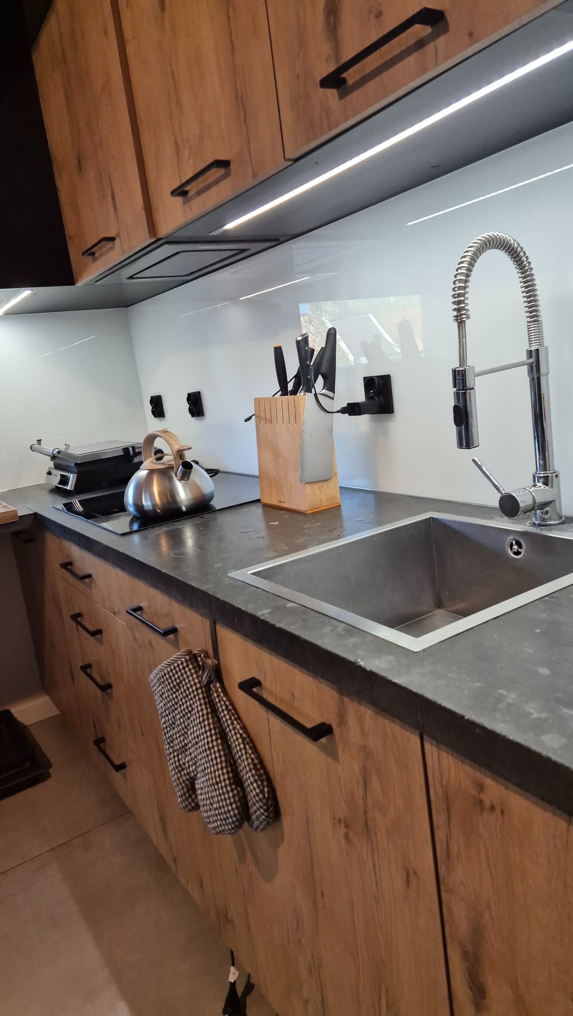 Nowoczesna kuchnia z drewnianymi szafkami, stalowym zlewem, czajnikiem i listwą nożową na blacie, z oświetleniem LED pod szafkami górnymi.