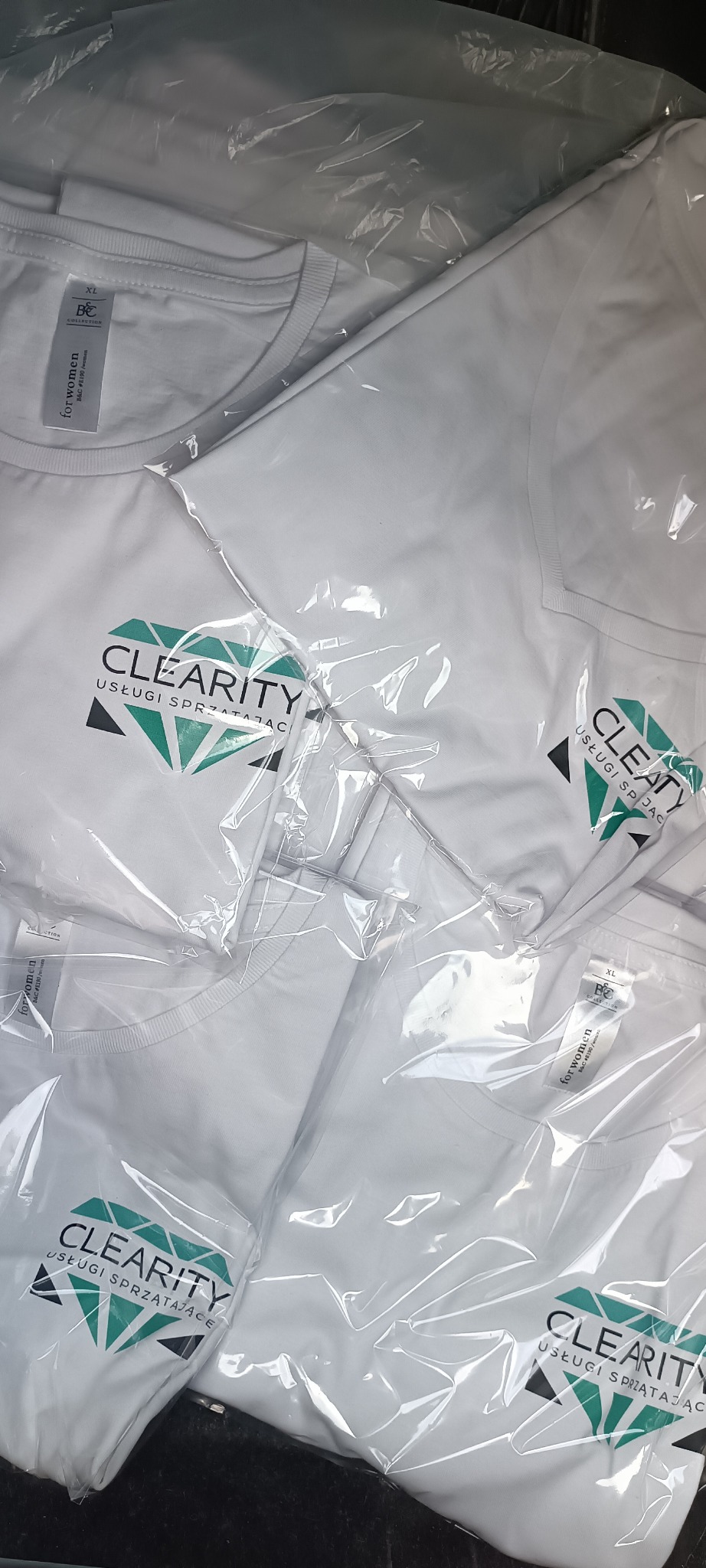 Stos białych koszulek z nadrukowanym logo firmy sprzątającej 'CLEARITY' w przezroczystych foliowych opakowaniach.