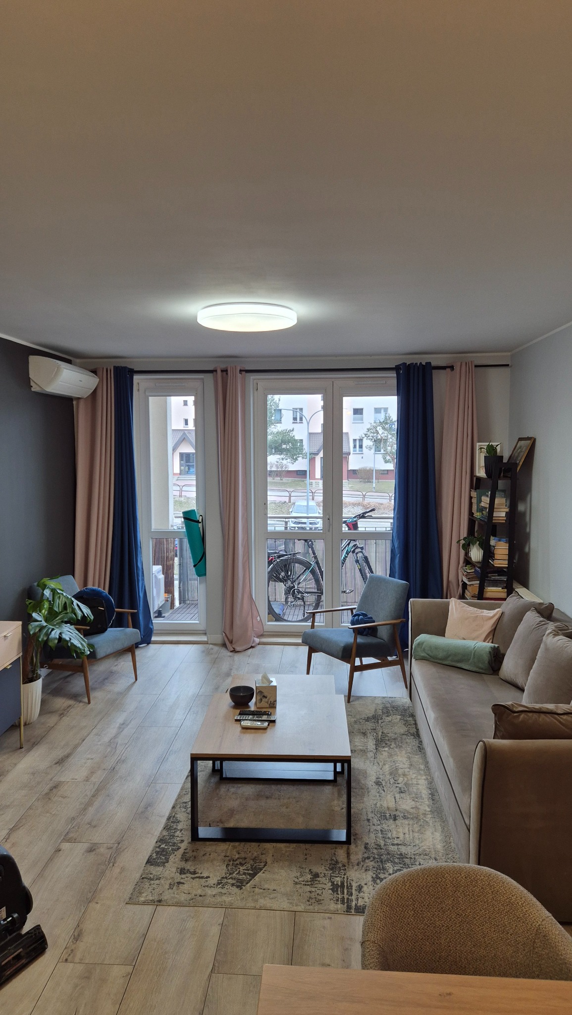 Ustawny salon z drewnianą podłogą, szarą sofą, dwoma fotelami i stolikiem kawowym na wzorzystym dywanie. Przez balkon widoczny rower. Ściany w odcieniach szarości i beżu, okna z zasłonami w kolorze...