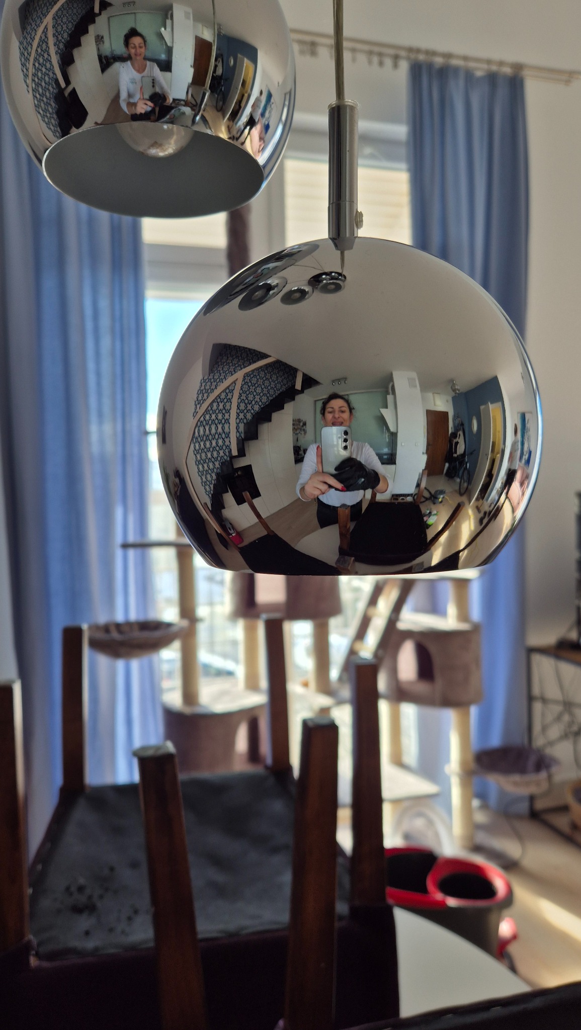 Odbicie wnętrza mieszkania w metalowej lampie wiszącej, widoczna osoba w rękawicach robiąca zdjęcie telefonem, w tle drapak dla kota i krzesło