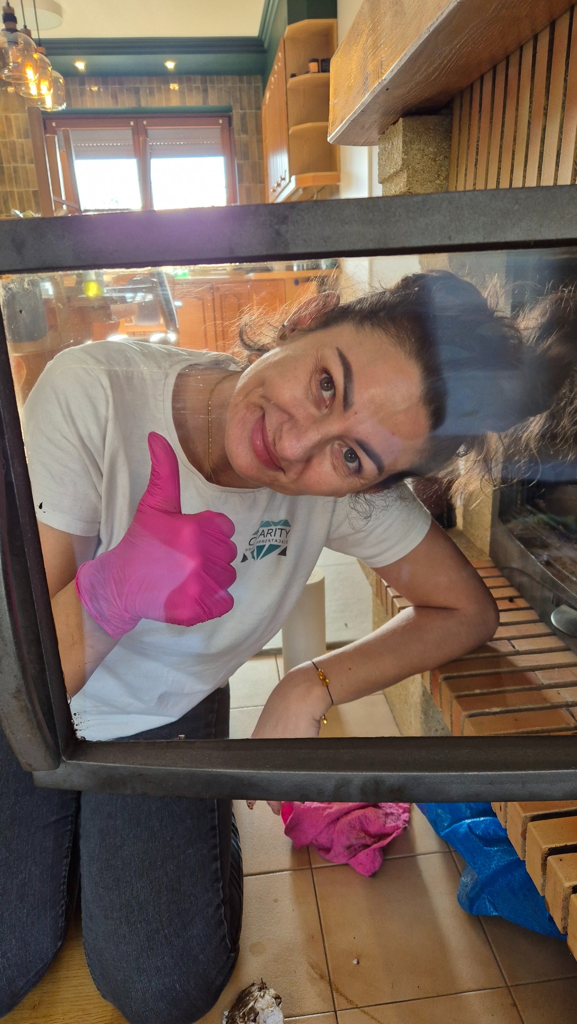 Uśmiechnięta kobieta w różowych rękawicach, ubrana w białą koszulkę z logo firmy sprzątającej, daje kciuk w górę, patrząc przez otwarte drzwiczki pieca kominkowego, widoczne wnętrze kuchni...