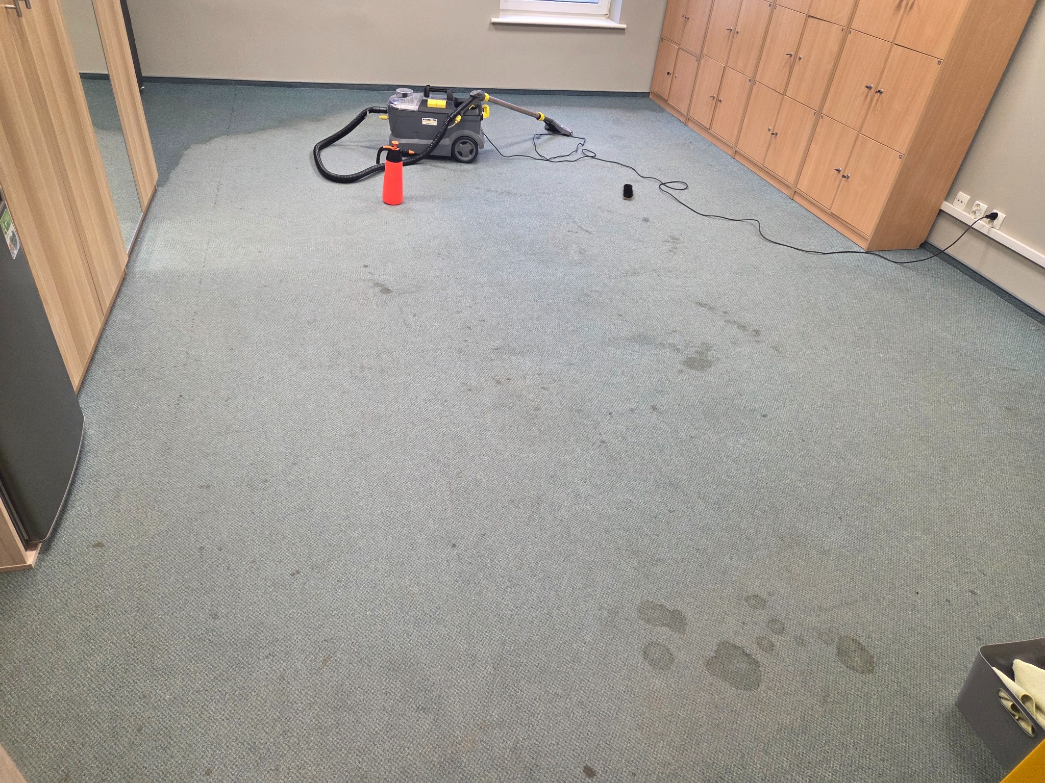 Szaro-niebieski dywan biurowy z widocznymi plamami, czyszczony za pomocą odkurzacza piorącego Karcher z czerwonym spryskiwaczem obok, w tle rząd szafek pracowniczych.