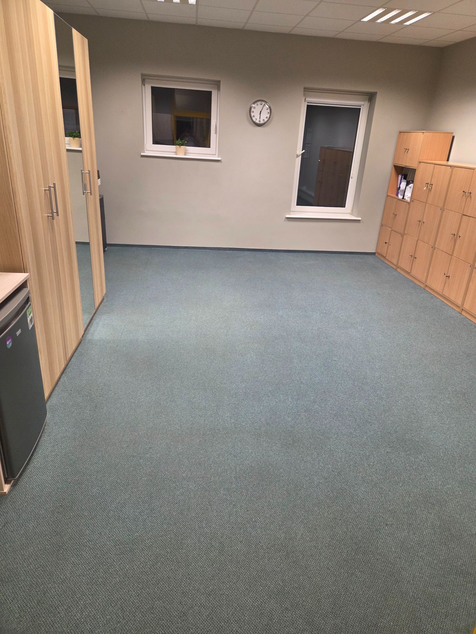 Pusta przestrzeń biurowa z szaro-niebieską wykładziną dywanową, jasnymi szafkami pracowniczymi, lustrzaną szafą, zegarem ściennym i dwoma oknami z roślinami w doniczkach na parapetach.