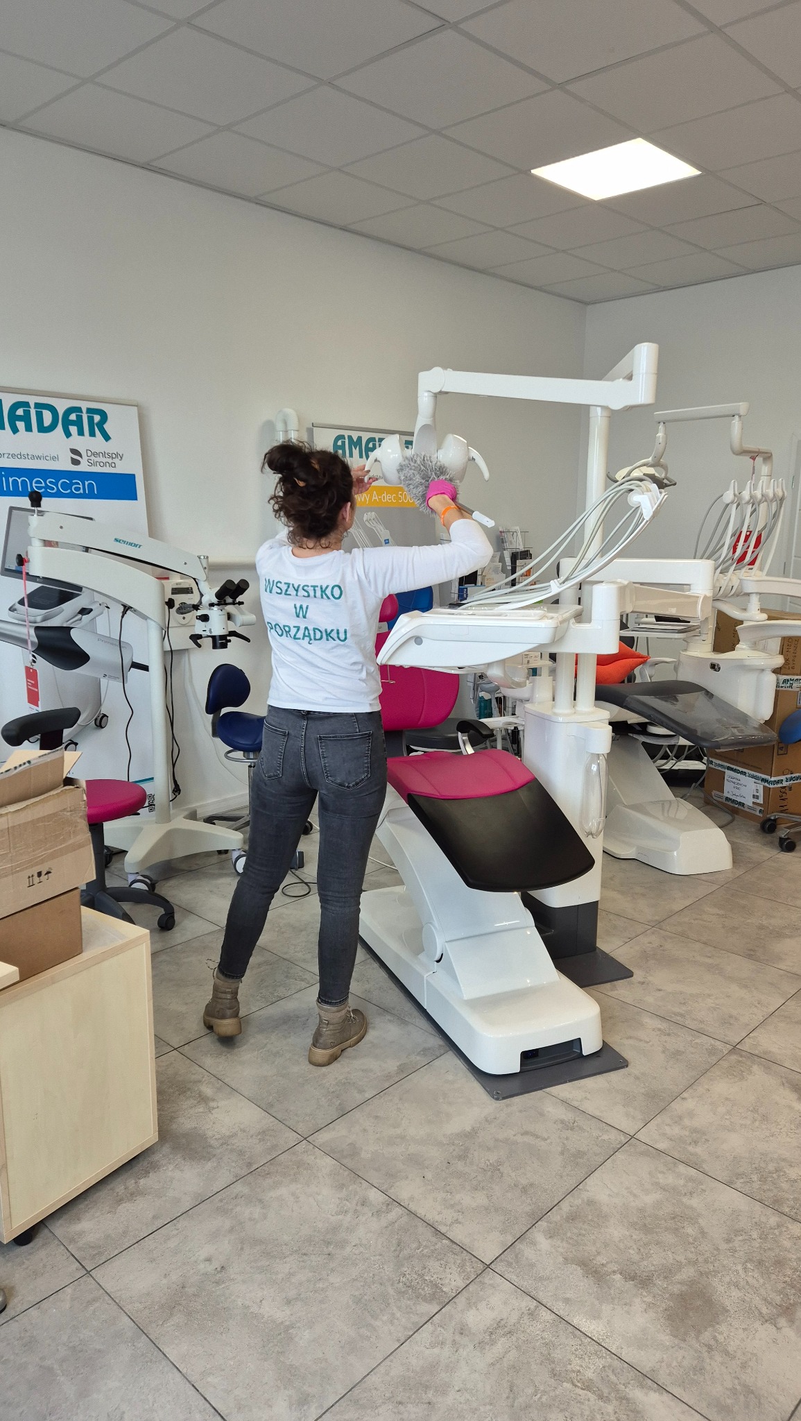 Kobieta w koszulce z napisem 'Wszystko w porządku' czyści za pomocą miotełki do kurzu biały unit stomatologiczny w gabinecie dentystycznym.