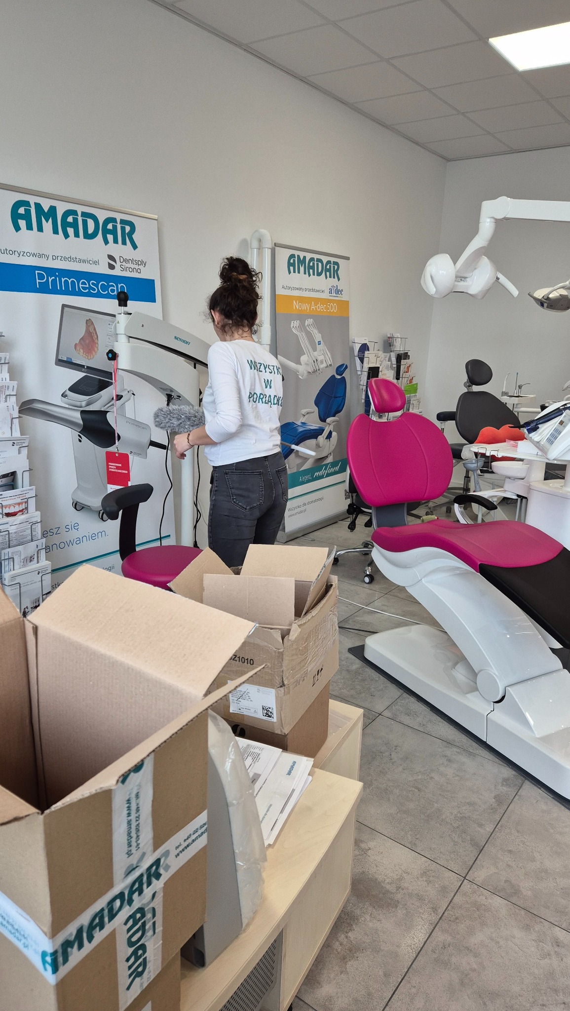 Kobieta czyści szczotką do kurzu skaner stomatologiczny Primescan w gabinecie dentystycznym, obok kartony z logo Amadar i fotel dentystyczny w kolorze różowym.