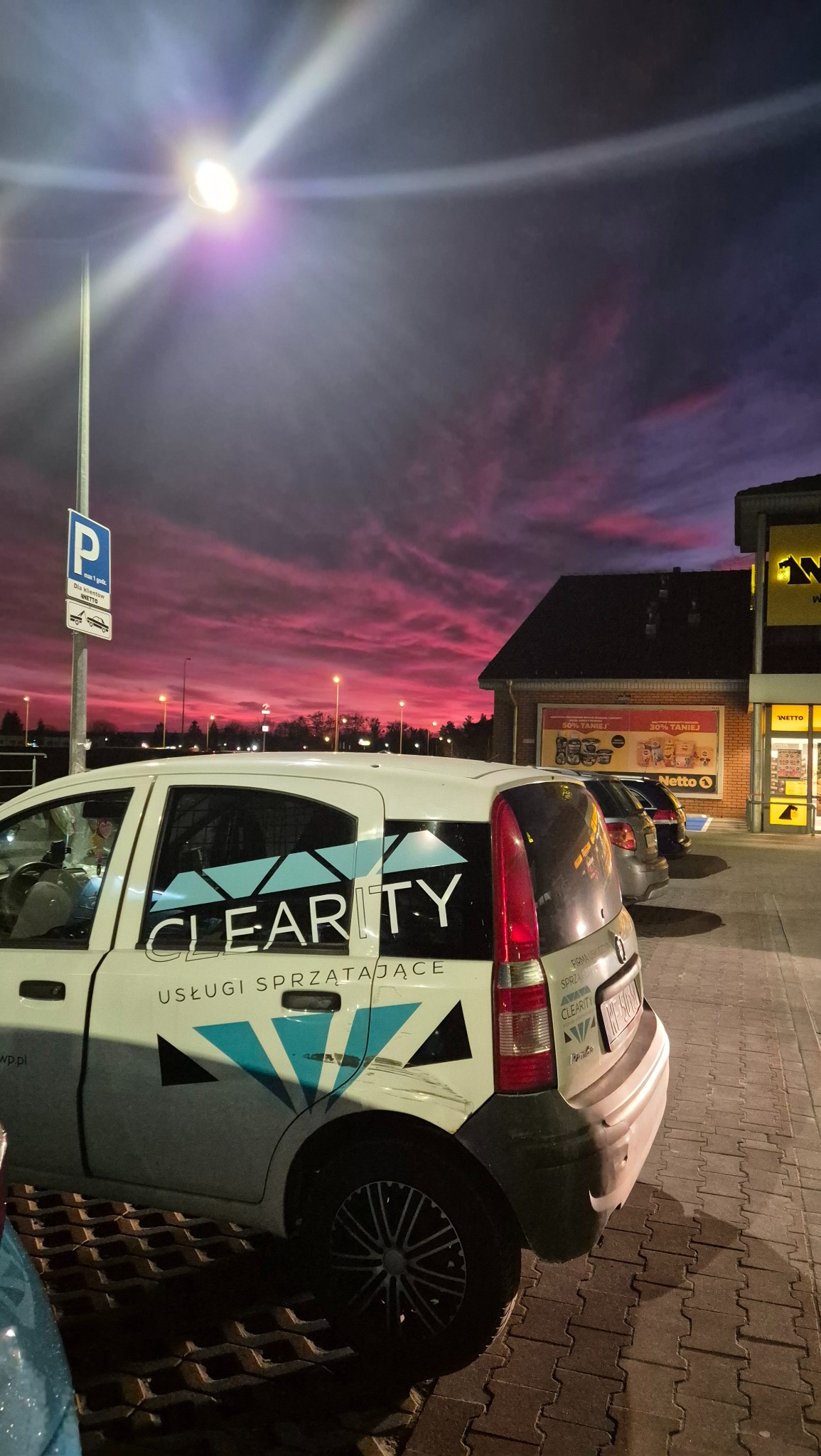 Samochód firmy sprzątającej Clarity zaparkowany przed sklepem Netto o zmierzchu, z widocznym logo i hasłem 'Usługi Sprzątające' na karoserii na tle intensywnie różowego nieba.