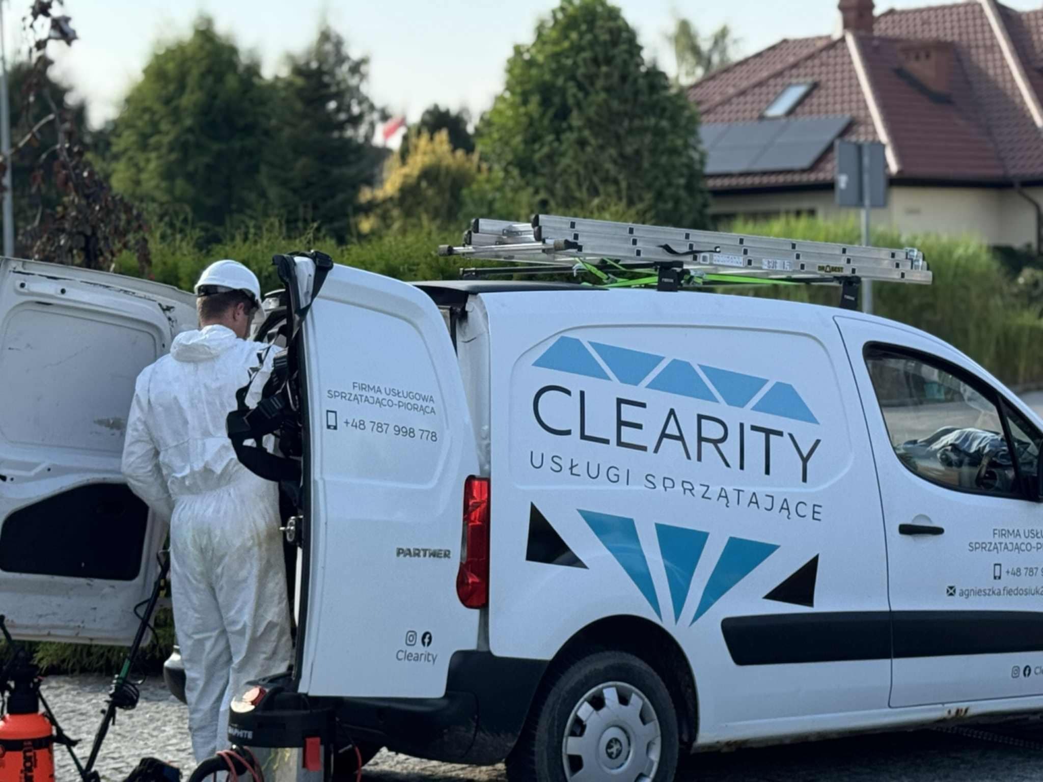Pracownik w kombinezonie przy samochodzie firmy sprzątającej 'Clearity' w Gdańsku. Widoczne drabiny na dachu i sprzęt do czyszczenia.