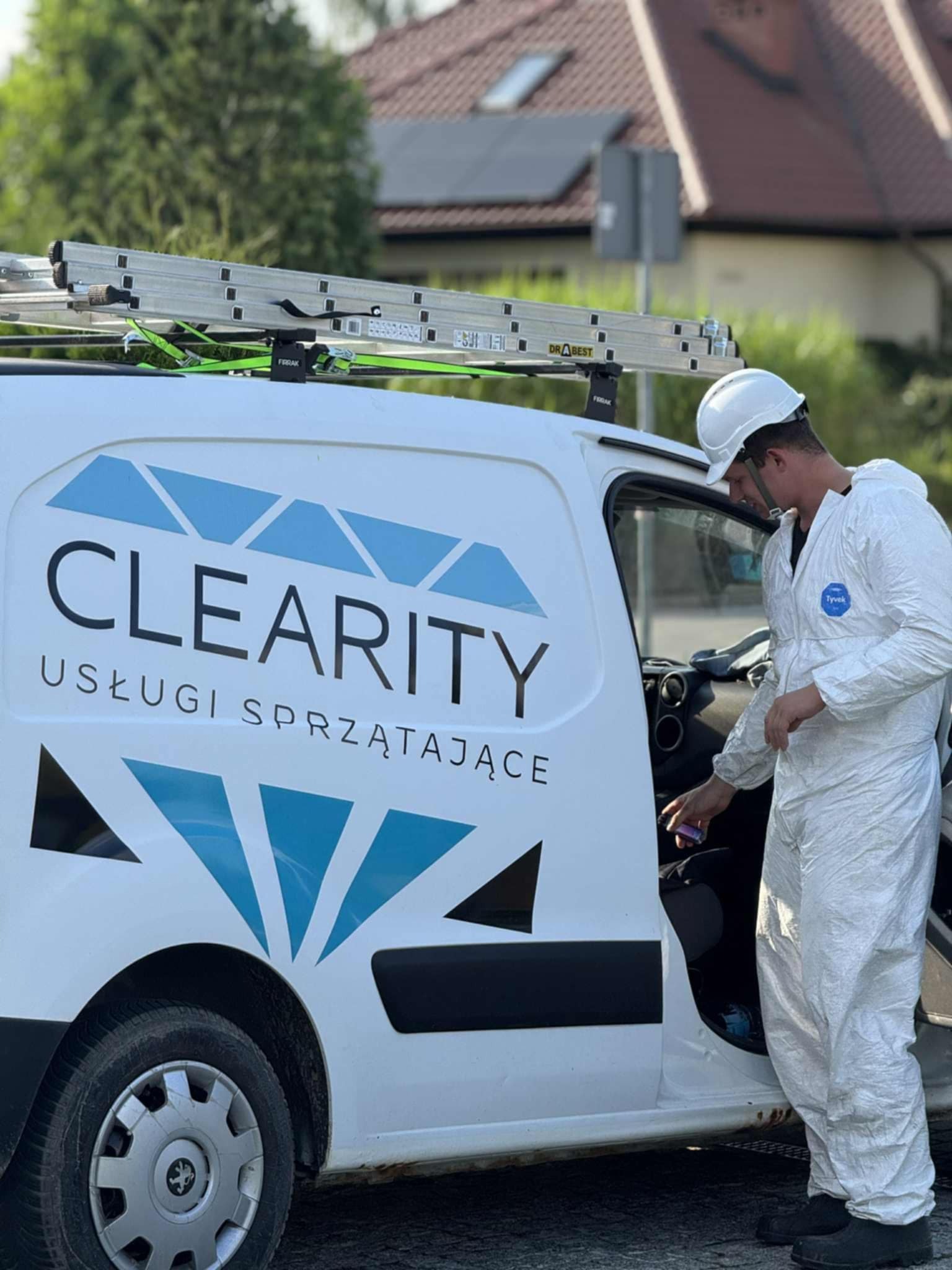 Pracownik w kombinezonie i kasku sięga do wnętrza białego busa z logo firmy sprzątającej. Na dachu drabina. Usługi sprzątające.