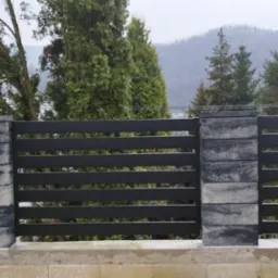 Nowoczesne, poziome ogrodzenie panelowe z ciemnoszarego metalu, wsparte na słupkach z szarej kostki betonowej, na tle zalesionego wzgórza i budynku z brązowym dachem.