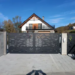 Nowoczesna, przesuwna brama wjazdowa z metalu w kolorze antracytowym, z laserowo wycinanym wzorem drzewa, na tle domu z balkonem i błękitnego nieba.