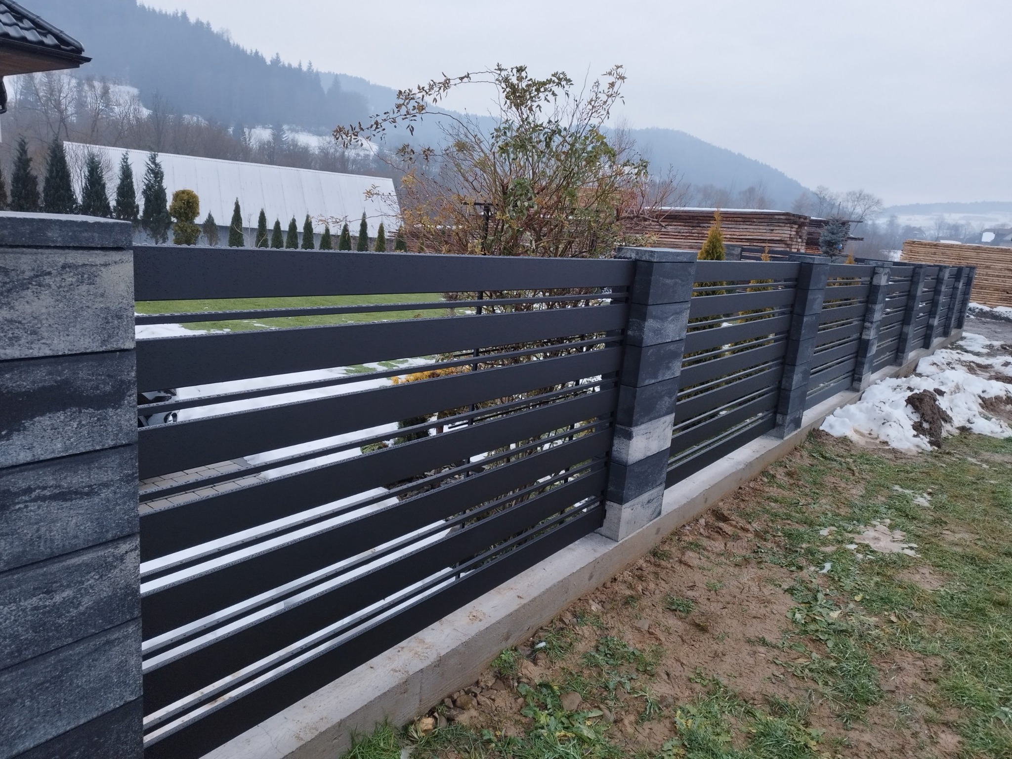 Nowoczesne ogrodzenie panelowe z szarymi betonowymi słupkami i poziomymi, czarnymi przęsłami, widok na górski krajobraz w pochmurny dzień.