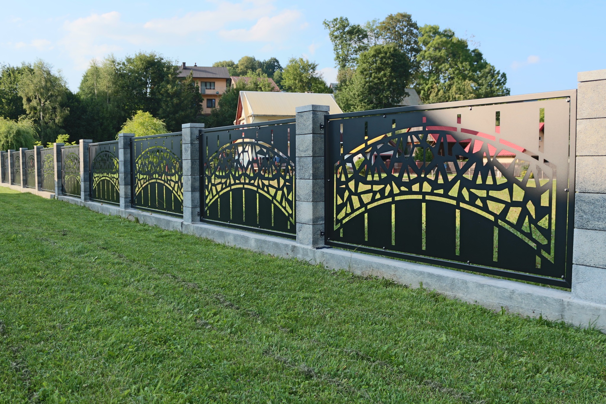 Długi, czarny, metalowy płot z ażurowym wzorem, osadzony na betonowej podmurówce i wsparty na kamiennych słupkach, rozciągający się wzdłuż trawnika przed domem.