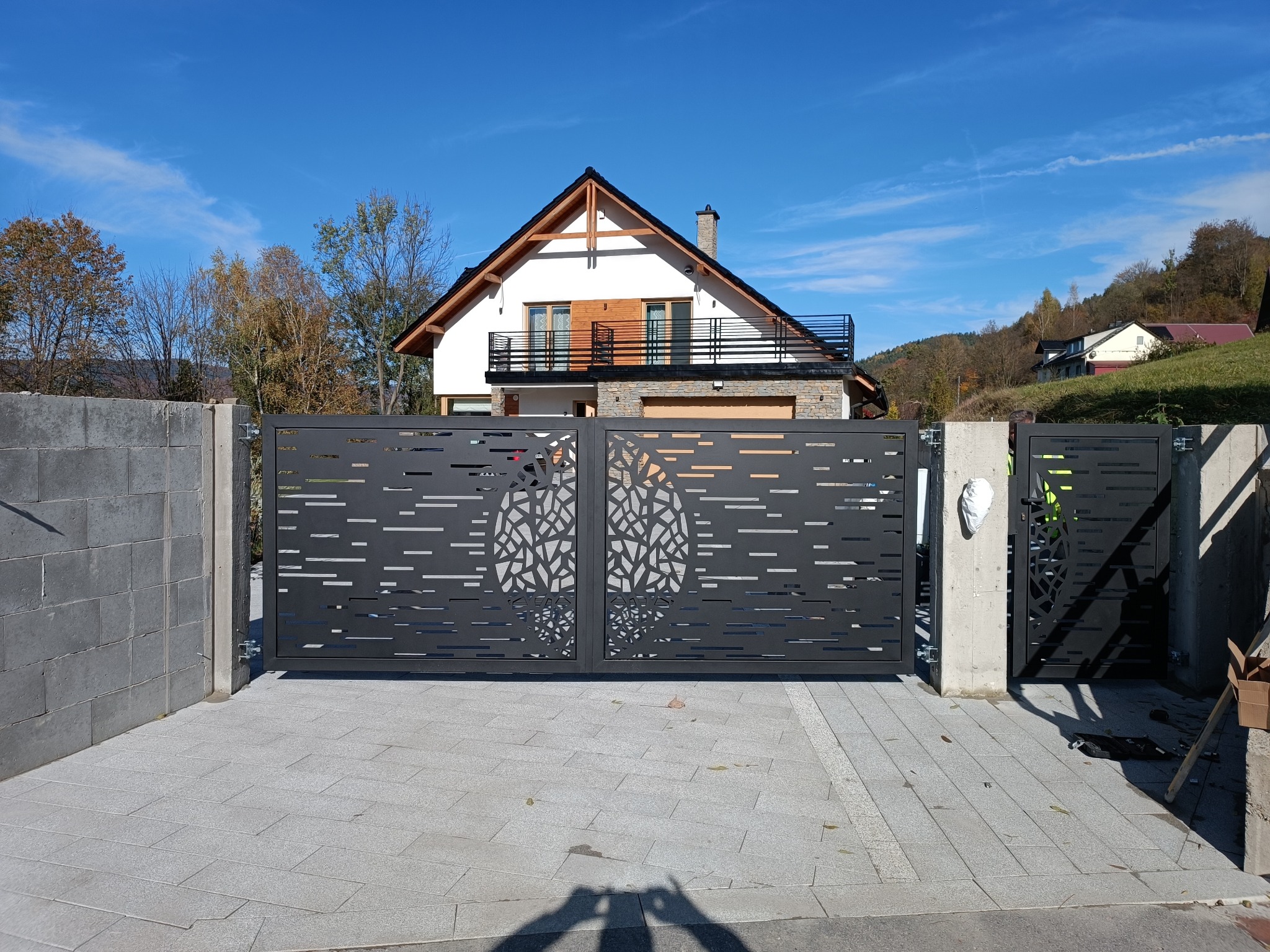 Nowoczesna, przesuwna brama w Tokarni z dekoracyjnym motywem drzewa, zamontowana na betonowych słupkach, na tle domu z balkonem i błękitnego nieba.