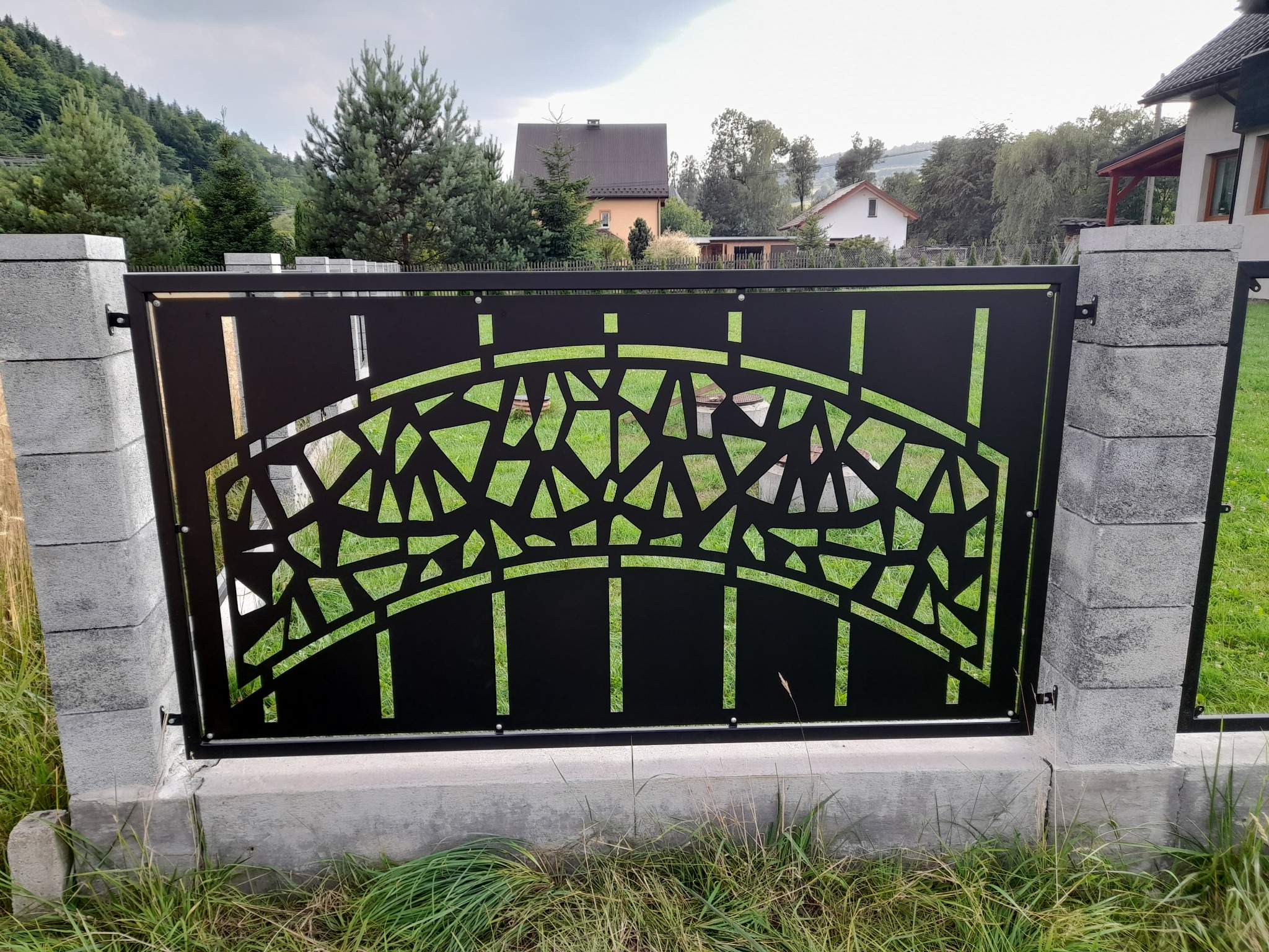 Czarny metalowy panel ogrodzeniowy z wycinanym wzorem geometrycznym, zamontowany między szarymi betonowymi słupkami, na tle zielonej trawy i zabudowań mieszkalnych.