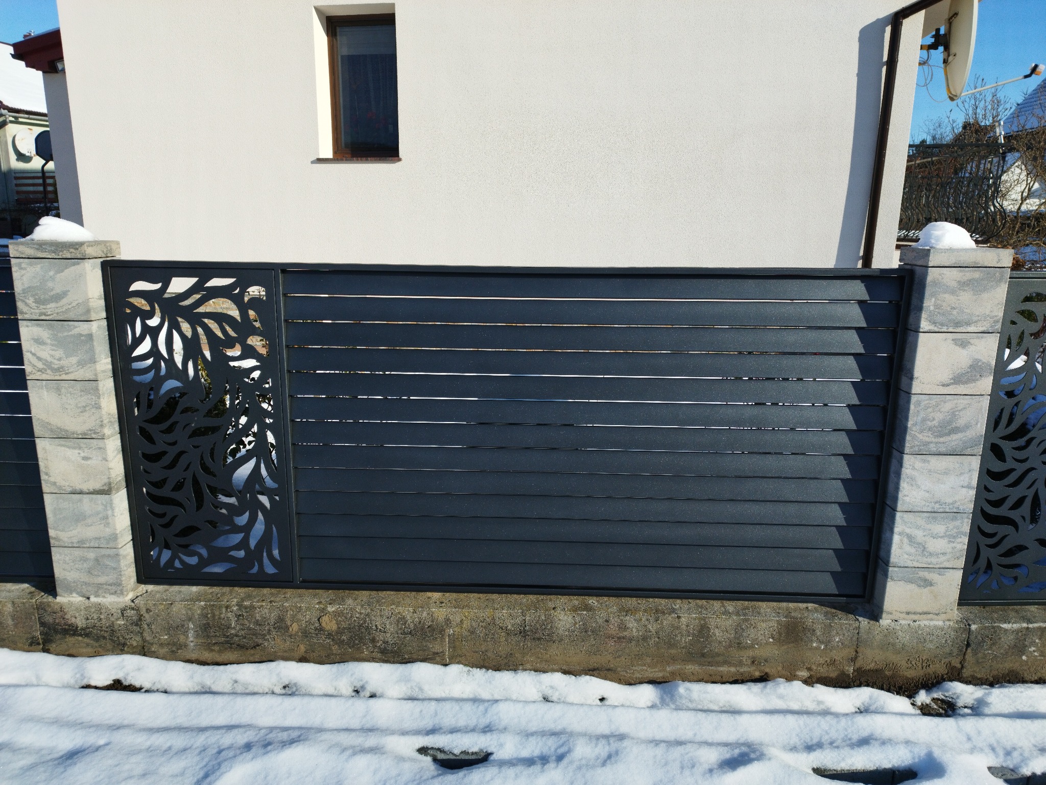 Nowoczesne ogrodzenie z poziomych paneli w kolorze antracytowym, z dekoracyjnymi elementami wycinanymi laserowo w motywy roślinne, osadzone na betonowej podmurówce i słupkach z szarej kostki,...