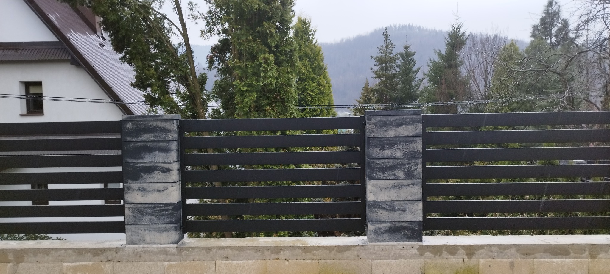 Nowoczesne, poziome ogrodzenie panelowe z ciemnoszarego metalu, wsparte na słupkach z szarej kostki betonowej, na tle zalesionego wzgórza i budynku z brązowym dachem.