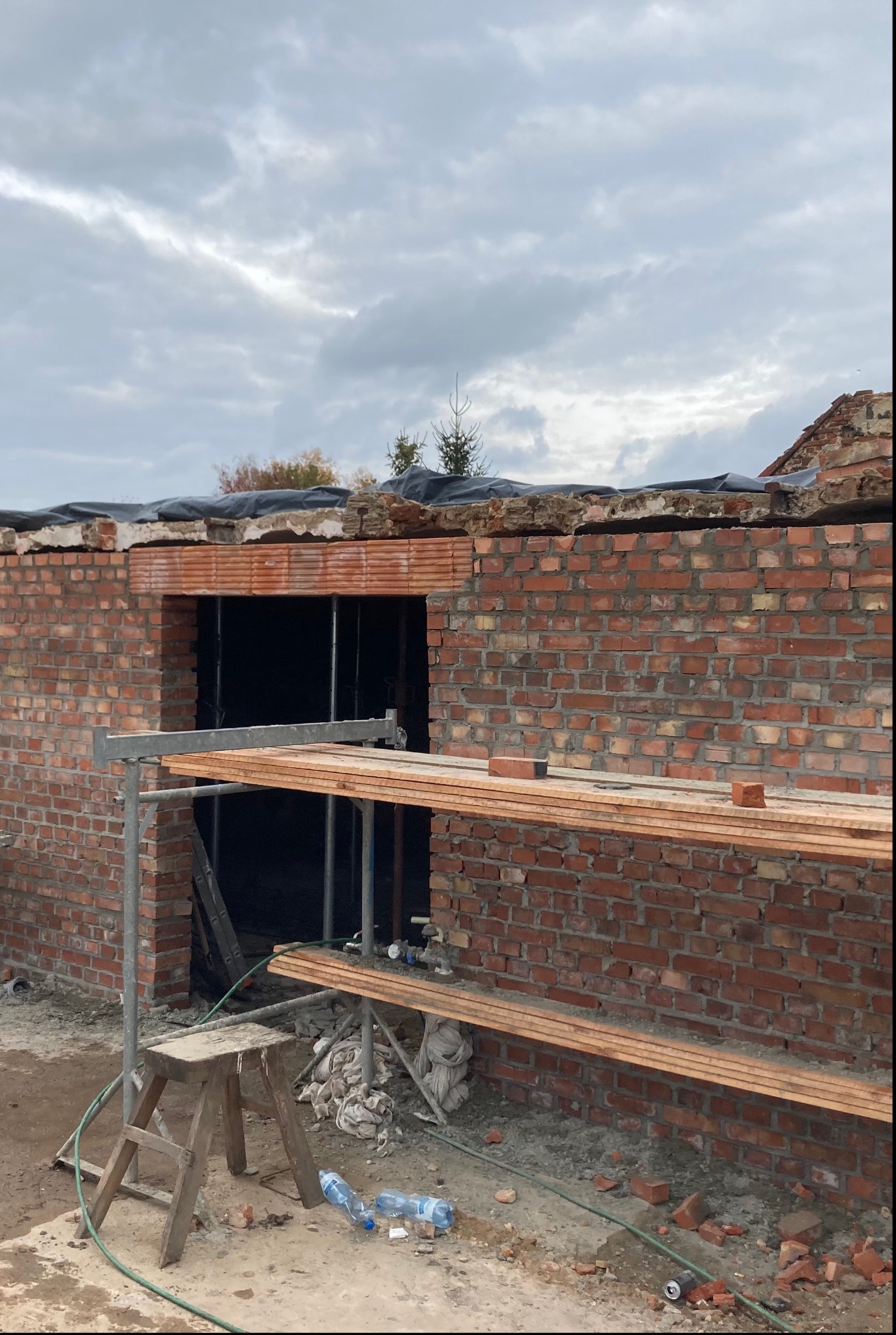 Budowa ściany z czerwonej cegły z widocznym otworem okiennym, rusztowaniem i drewnianym stołkiem na placu budowy w pochmurny dzień.