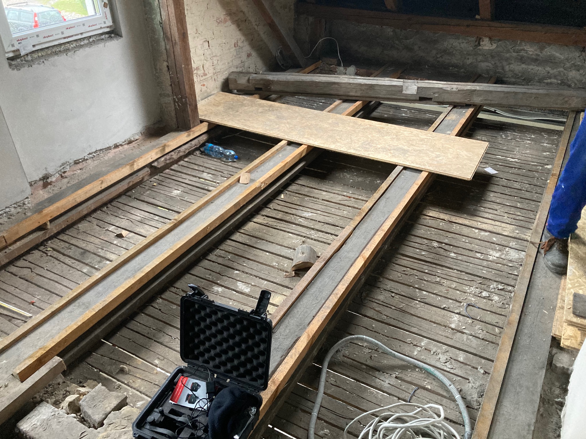 Stare, drewniane poddasze w trakcie remontu, widoczne odsłonięte belki stropowe, częściowo ułożona płyta OSB, butelka wody, skrzynka z narzędziami i fragment niebieskiej odzieży roboczej.