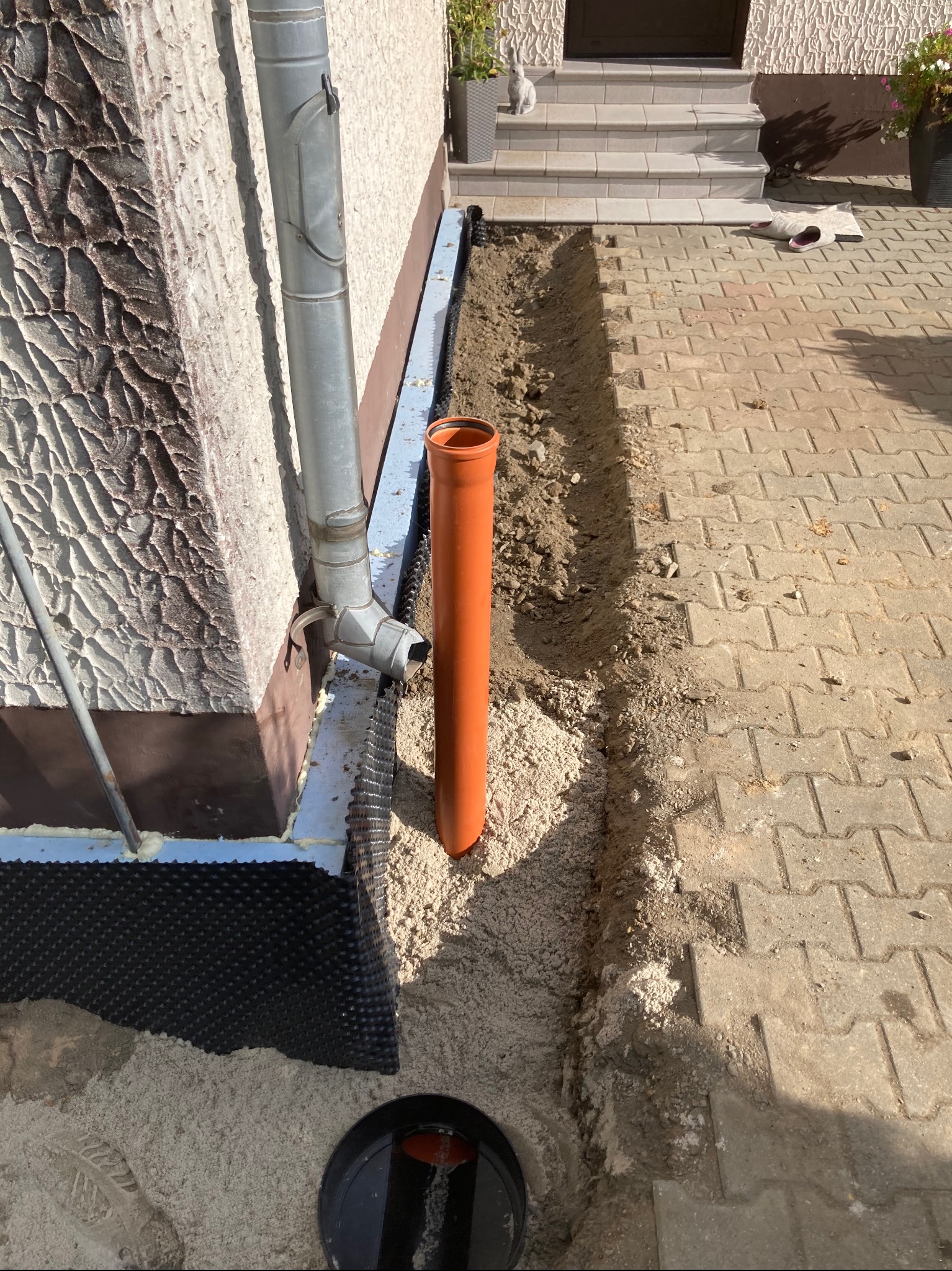 Fundament domu z izolacją pionową, rura drenarska i studzienka rewizyjna w wykopie, widoczna faktura elewacji i rynna spustowa.