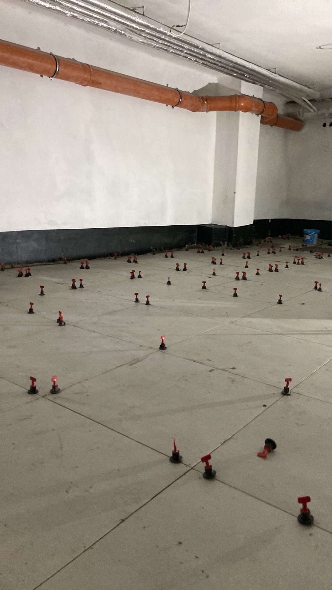 Szare płytki podłogowe w trakcie układania, z użyciem czerwonych klinów poziomujących, widoczne rury pod sufitem.