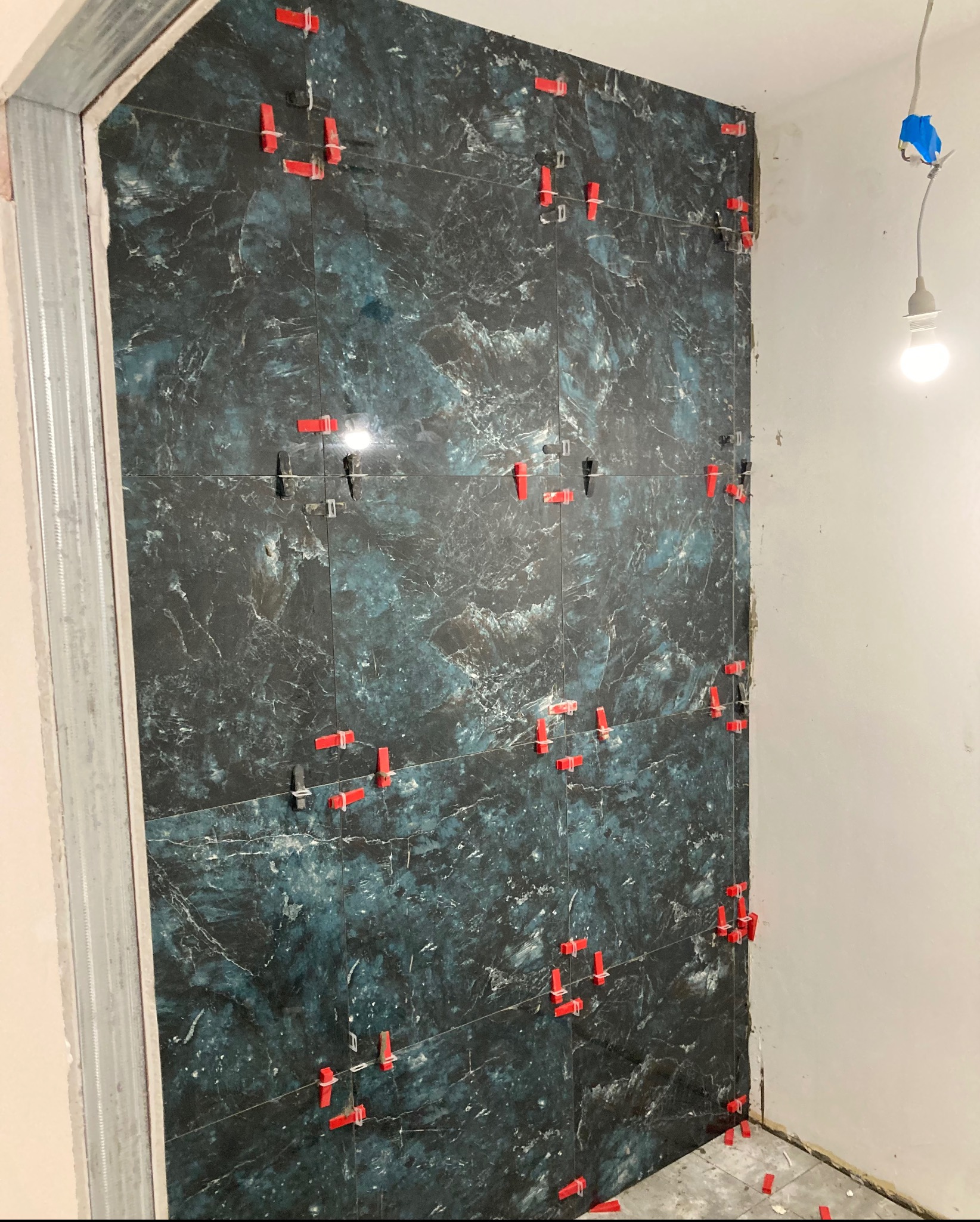 Ściana w trakcie układania płytek imitujących marmur, z widocznymi czerwonymi krzyżykami dystansowymi i ramą z profili stalowych przy otworze drzwiowym.