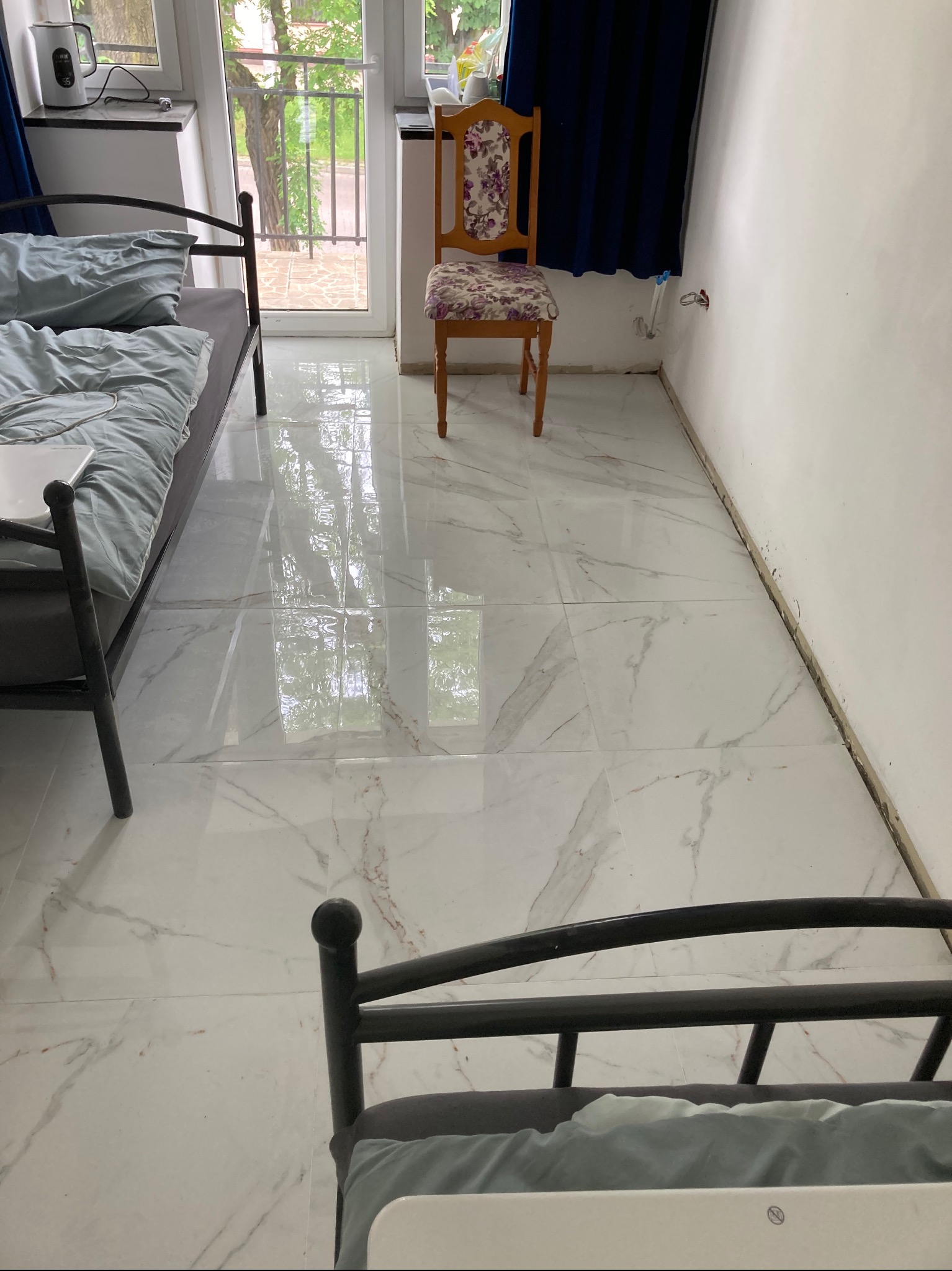 Pokój w trakcie remontu: położone białe płytki imitujące marmur, widoczne dwa łóżka z pościelą, krzesło z kwiatowym wzorem i odsłonięte kable w ścianie.