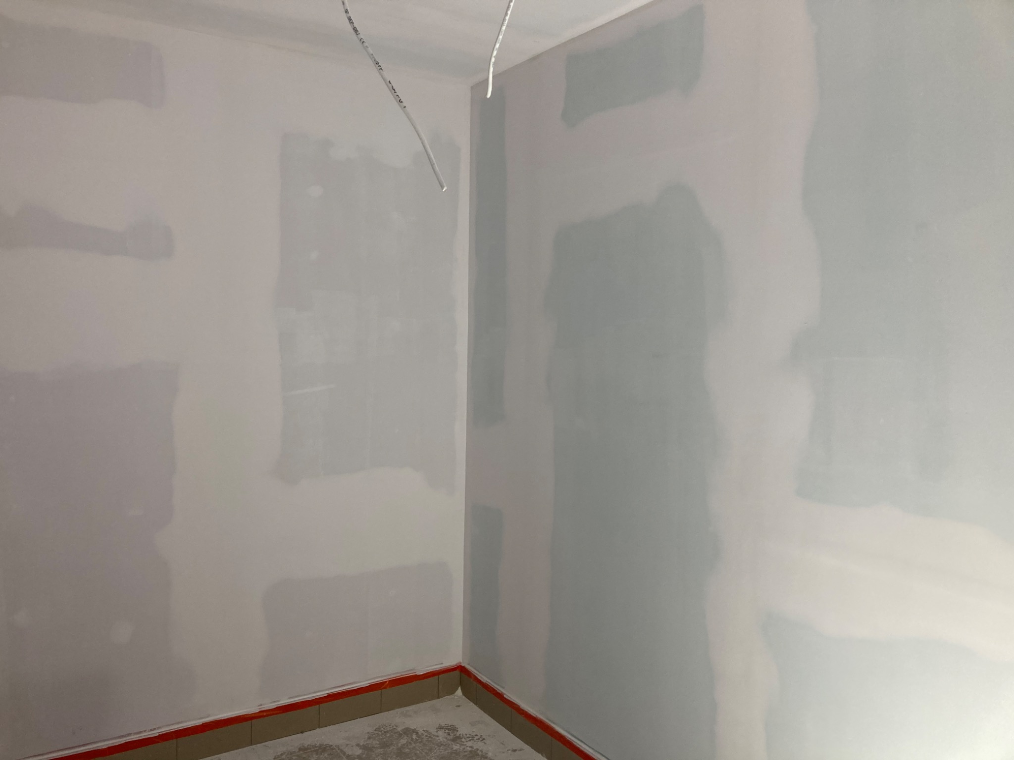 Ściany w pokoju po szpachlowaniu, przygotowane do malowania, z widocznymi przewodami elektrycznymi i pomarańczową taśmą zabezpieczającą przy podłodze.