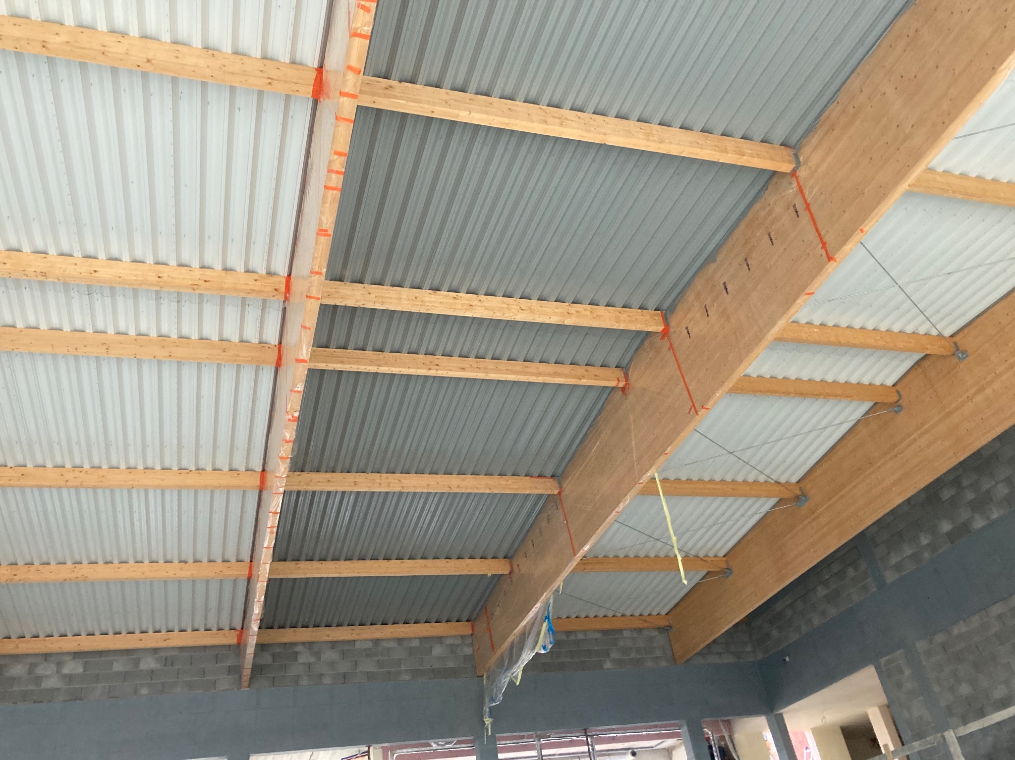 Wnętrze budynku w trakcie budowy, widoczna konstrukcja dachu z drewnianych belek i blachy trapezowej, zabezpieczona pomarańczową taśmą, ściany z szarego betonu komórkowego.