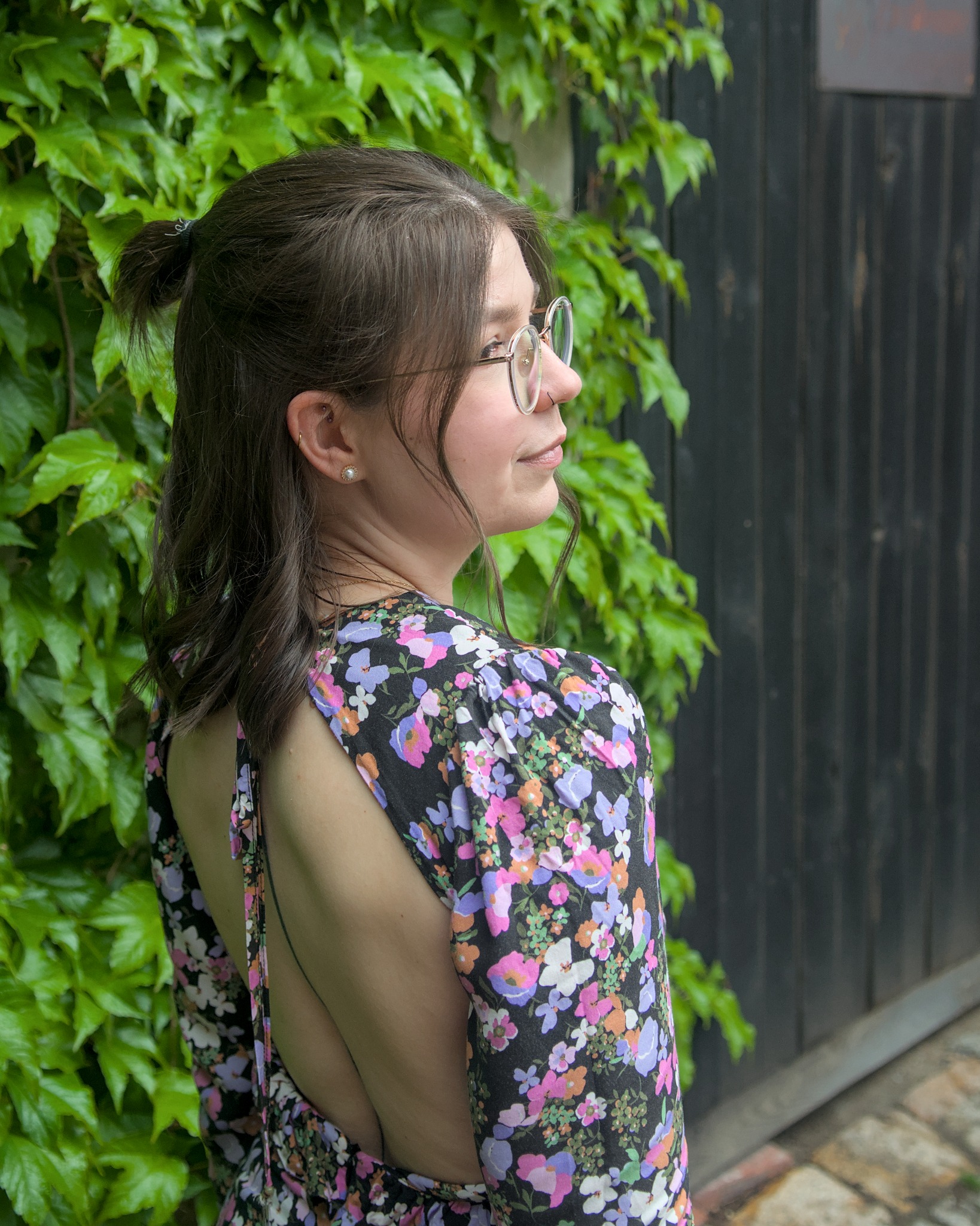 Portret kobiety w kwiecistej sukience, z odsłoniętymi plecami i okularach, stojącej na tle ściany z bluszczu i drewnianej ściany. Profil, naturalne światło.