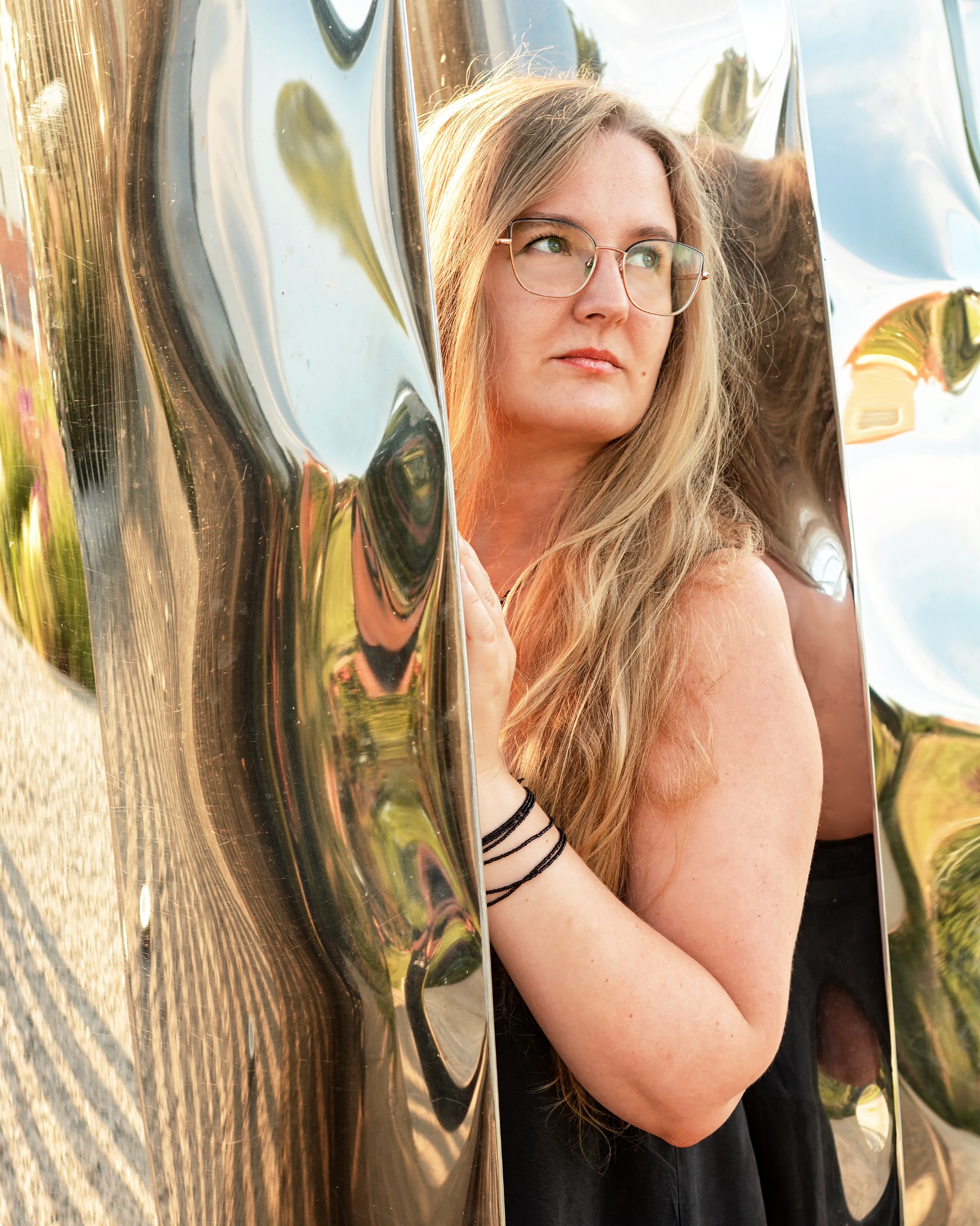 Portret kobiety w okularach, z blond włosami, pozującej obok lustrzanej, falistej powierzchni, która odbija otoczenie. Ujęcie w plenerze, w naturalnym świetle.