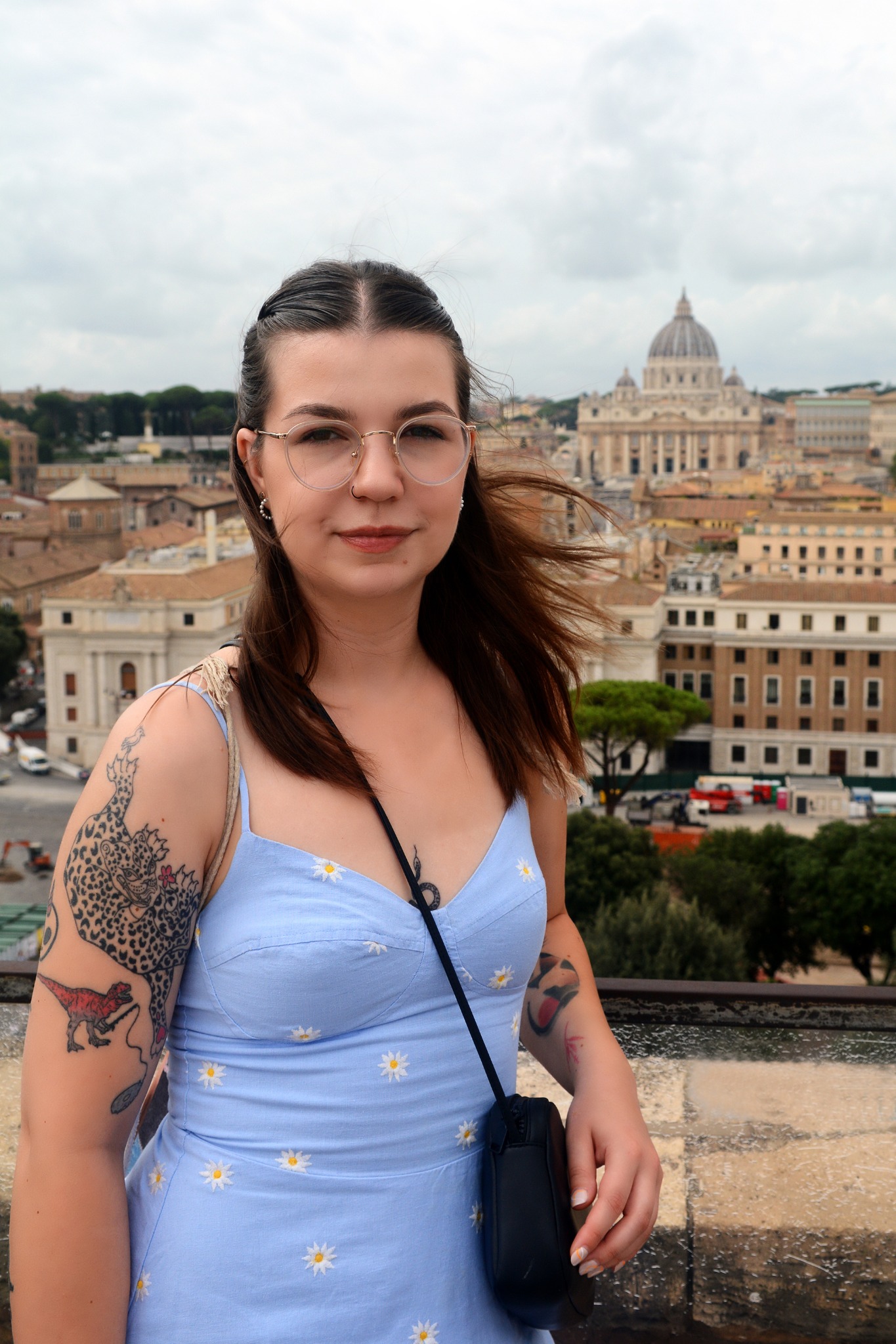 Portret kobiety w okularach na tle panoramy Rzymu z Bazyliką św. Piotra. Dziewczyna w sukience w kwiatki ma tatuaże na ramionach.