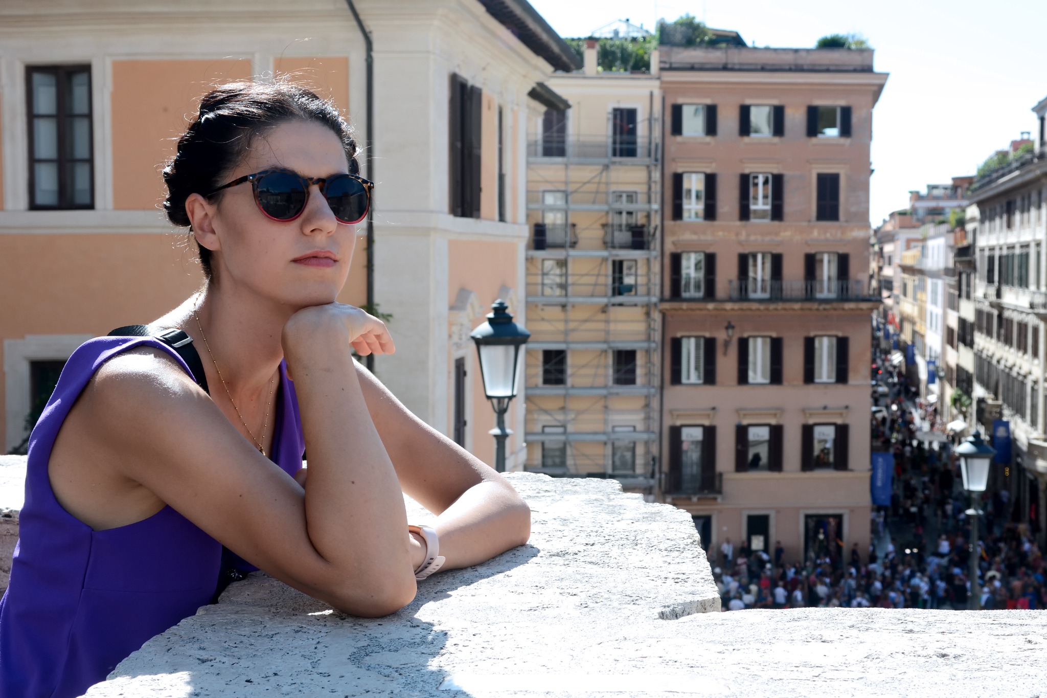 Kobieta w okularach przeciwsłonecznych opiera się o kamienny mur z widokiem na rzymską ulicę wypełnioną ludźmi i zabytkową architekturą.