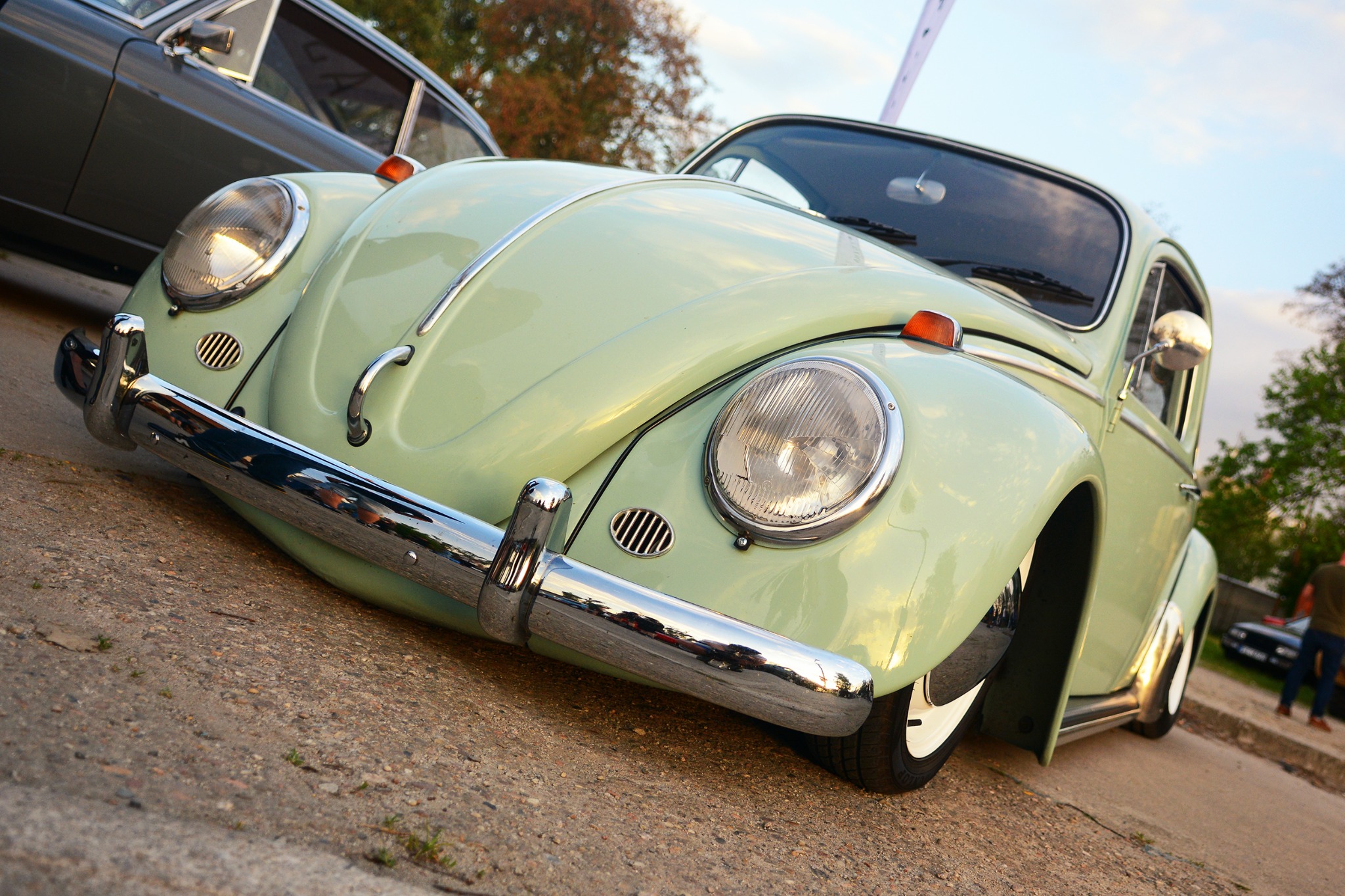 Nietypowe ujęcie od dołu na odrestaurowanego, jasnozielonego Volkswagena Garbusa z chromowanymi zderzakami, zaparkowanego na tle innych aut i zieleni.