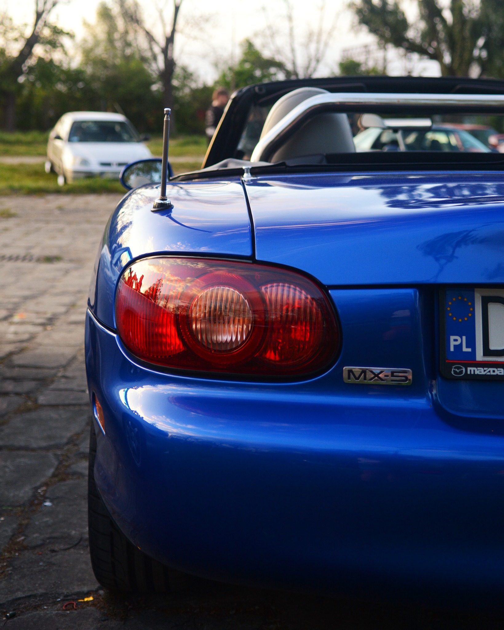 Błękitny kabriolet Mazda MX-5 z widocznymi tylnymi światłami i tablicą rejestracyjną PL, zaparkowany na brukowanej powierzchni w plenerze.