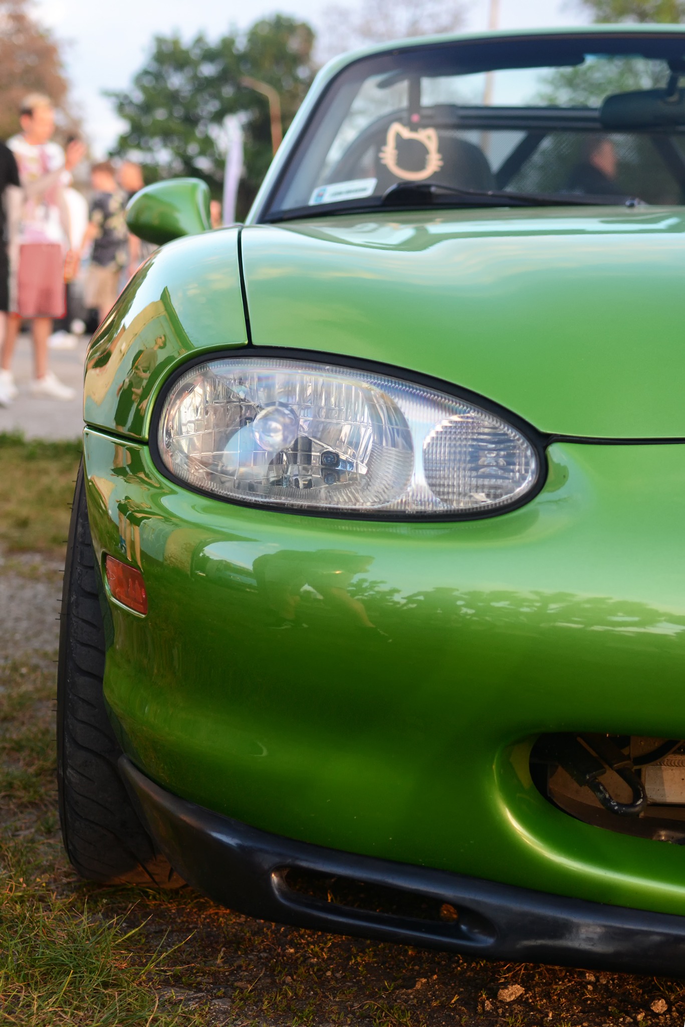 Zielony, tuningowany samochód Mazda MX-5 Miata z ozdobą Hello Kitty na desce rozdzielczej, sfotografowany z bliska, z widocznym reflektorem i fragmentem opony.
