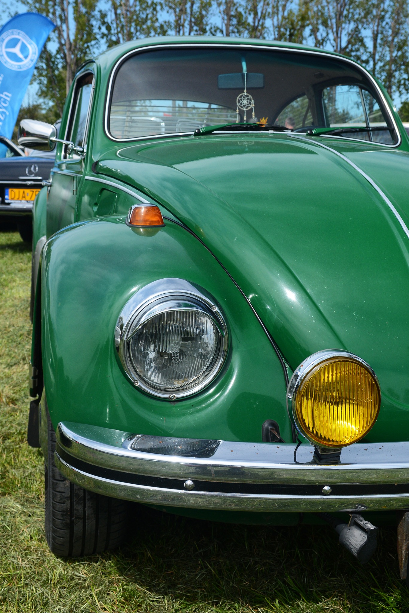 Zielony Volkswagen Garbus z chromowanym zderzakiem i żółtym reflektorem przeciwmgielnym, zaparkowany na trawie. Widoczny detal: łapacz snów na przedniej szybie.