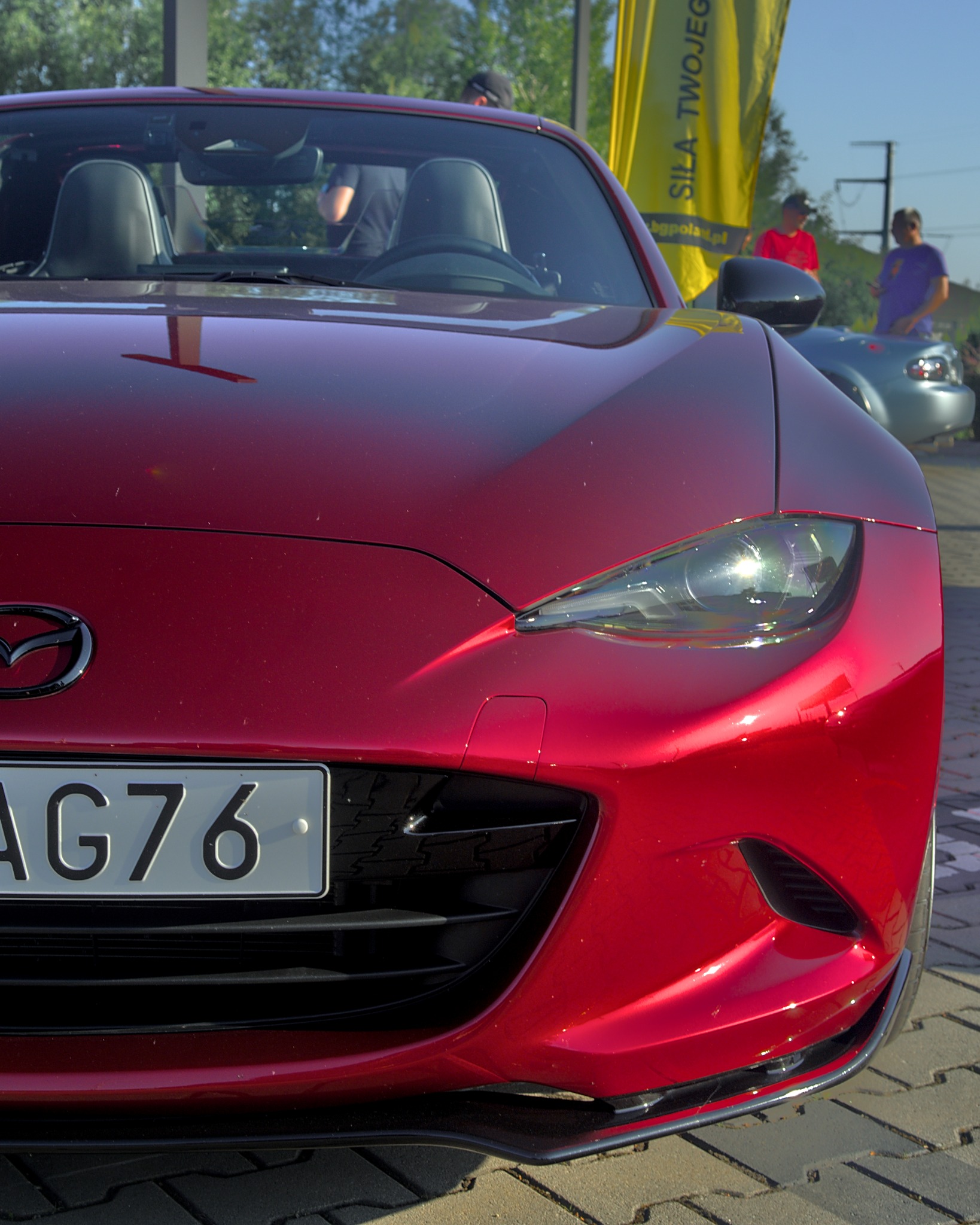Czerwona Mazda MX-5 z przodu, tablica AG76, na tle żółtego baneru i innych osób. Ostre światło podkreśla detale karoserii i reflektorów.